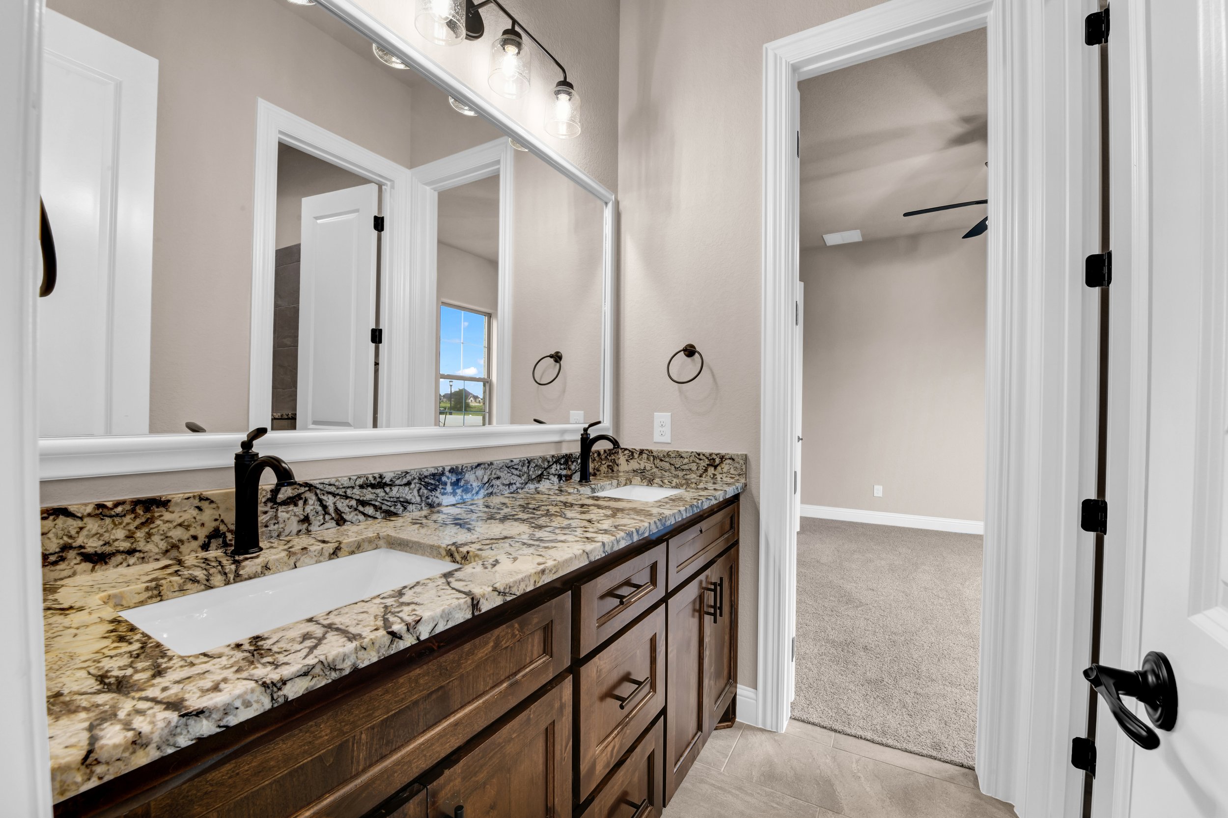 Bathroom with a granite countertop double sink vanity, large mirror, beige walls, and an open door leading to a room with beige carpet and a ceiling fan.