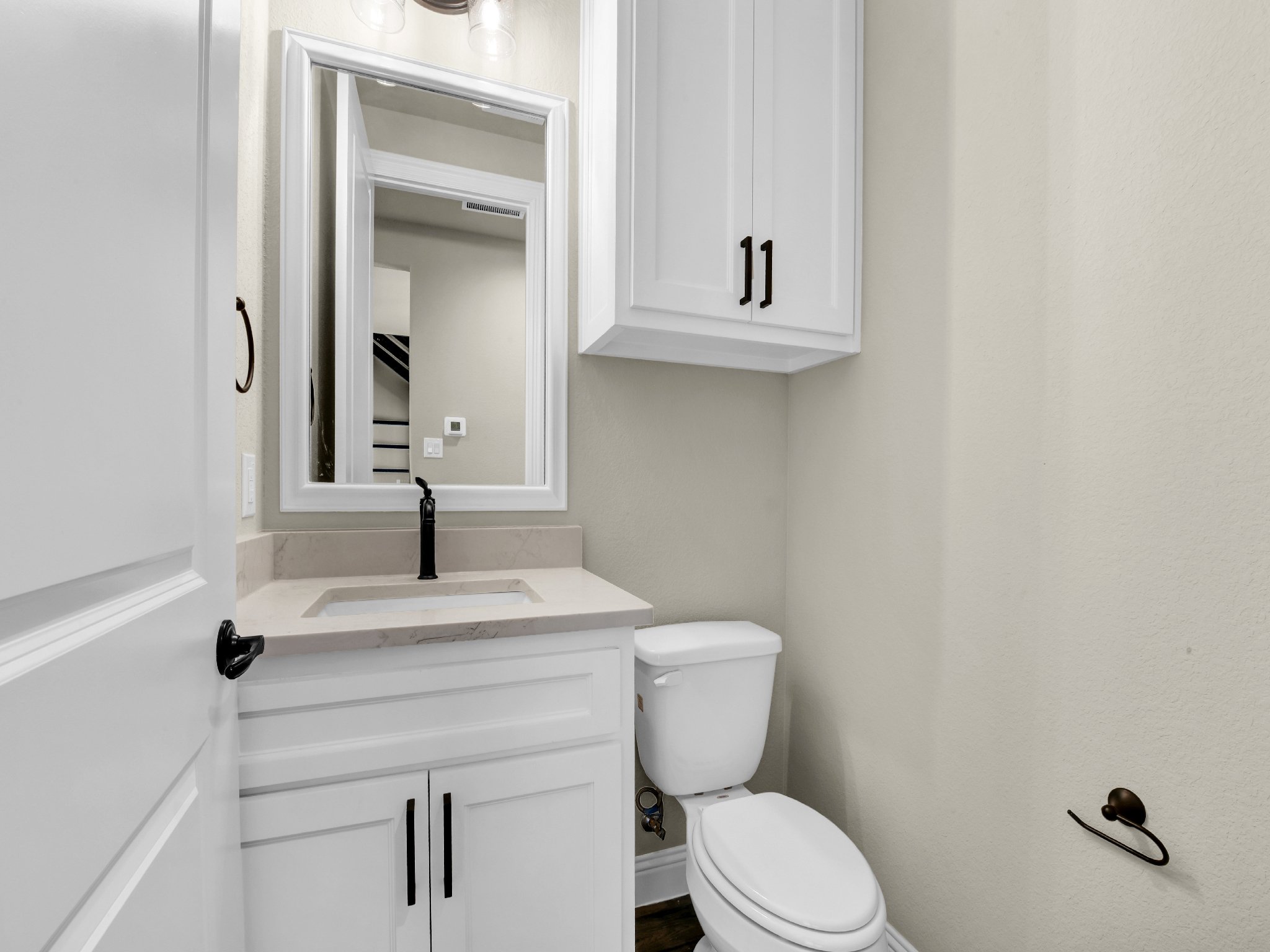 Small bathroom with white cabinetry, a mirror above a small sink, a white toilet, a wall-mounted cabinet, and a bronze toilet paper holder.