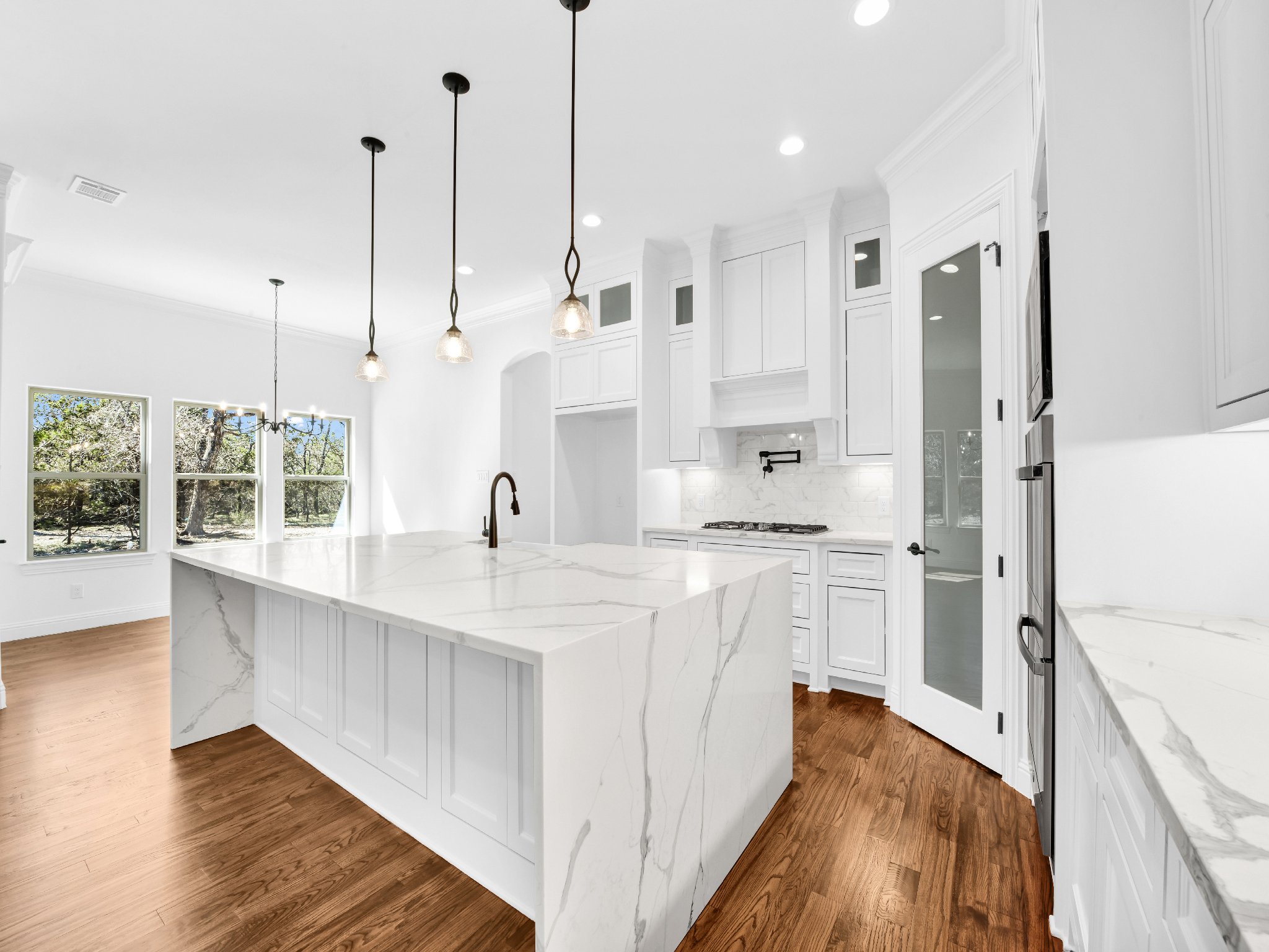 Modern white kitchen with marble countertops, white cabinets, and hardwood floors, illuminated by pendant and recessed lighting, with a bright outdoor view through large windows.