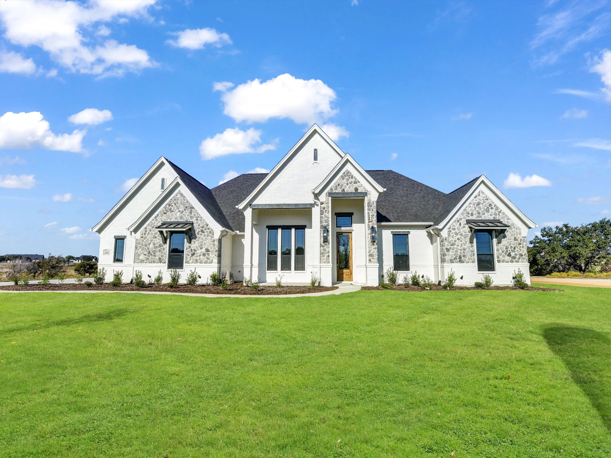 White house with stone accents, surrounded by a green lawn under a blue sky with scattered clouds.
