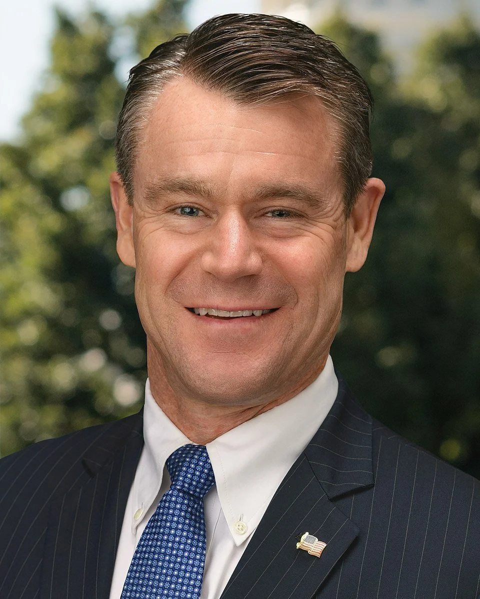 Man wearing a suit and tie with a small American flag pin, outdoors with greenery in the background.