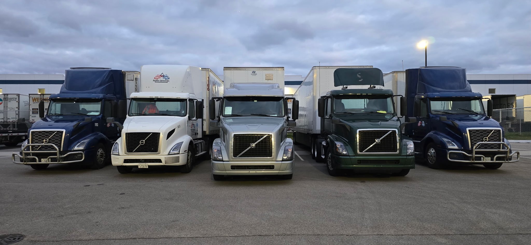 Four Partner Carriers Inc. semi trucks parked side by side at a truck yard with trailers attached, featuring different colors including blue, white, silver, and green.