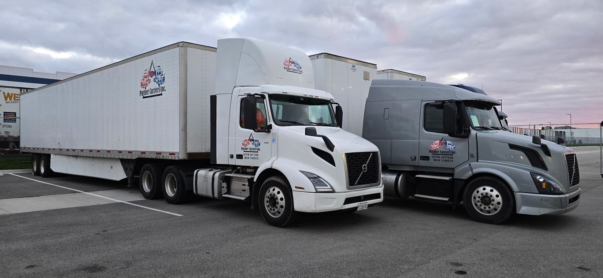 Two semi-trucks parked side by side, one white and one gray, both with the logo "Partner Carriers Inc." and DOT details on the doors, in a parking area under a cloudy sky.