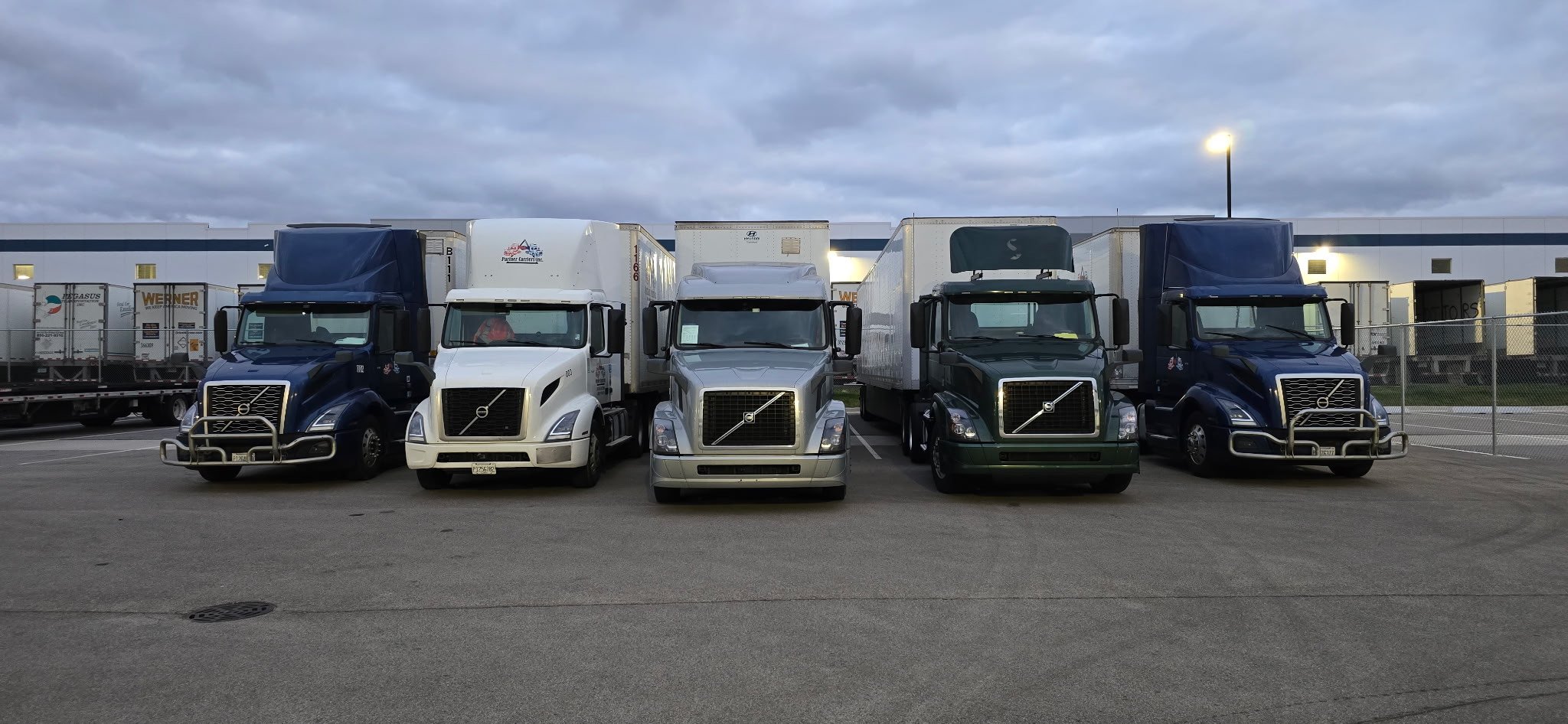 Four Partner Carriers Inc. Volvo semi-trucks parked in a row at a loading dock, with a cloudy sky overhead. The trucks have different colors: two blue, one white, and one green.