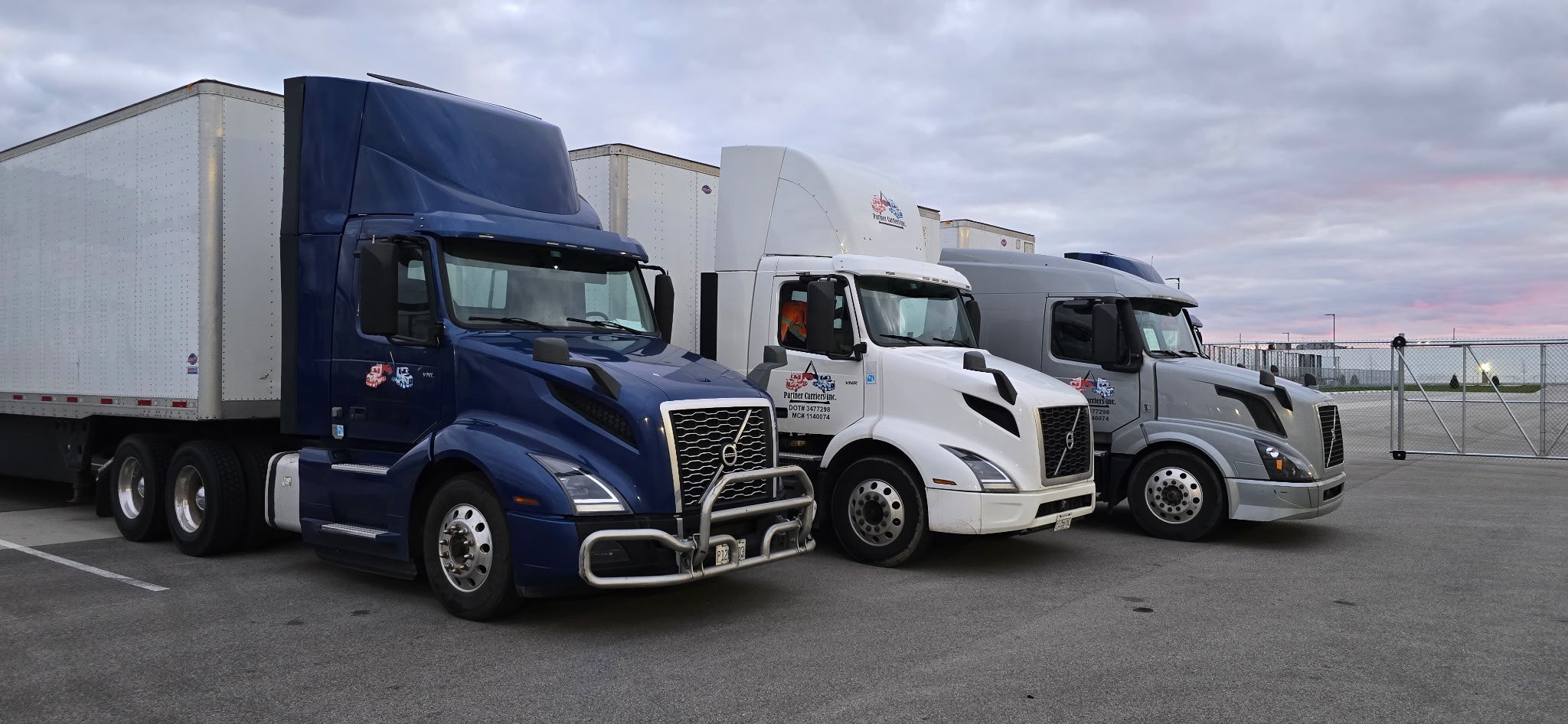 Three Partner Carriers Inc. semi-trucks parked in a lot under a cloudy sky, with different colors including blue, white, and gray.