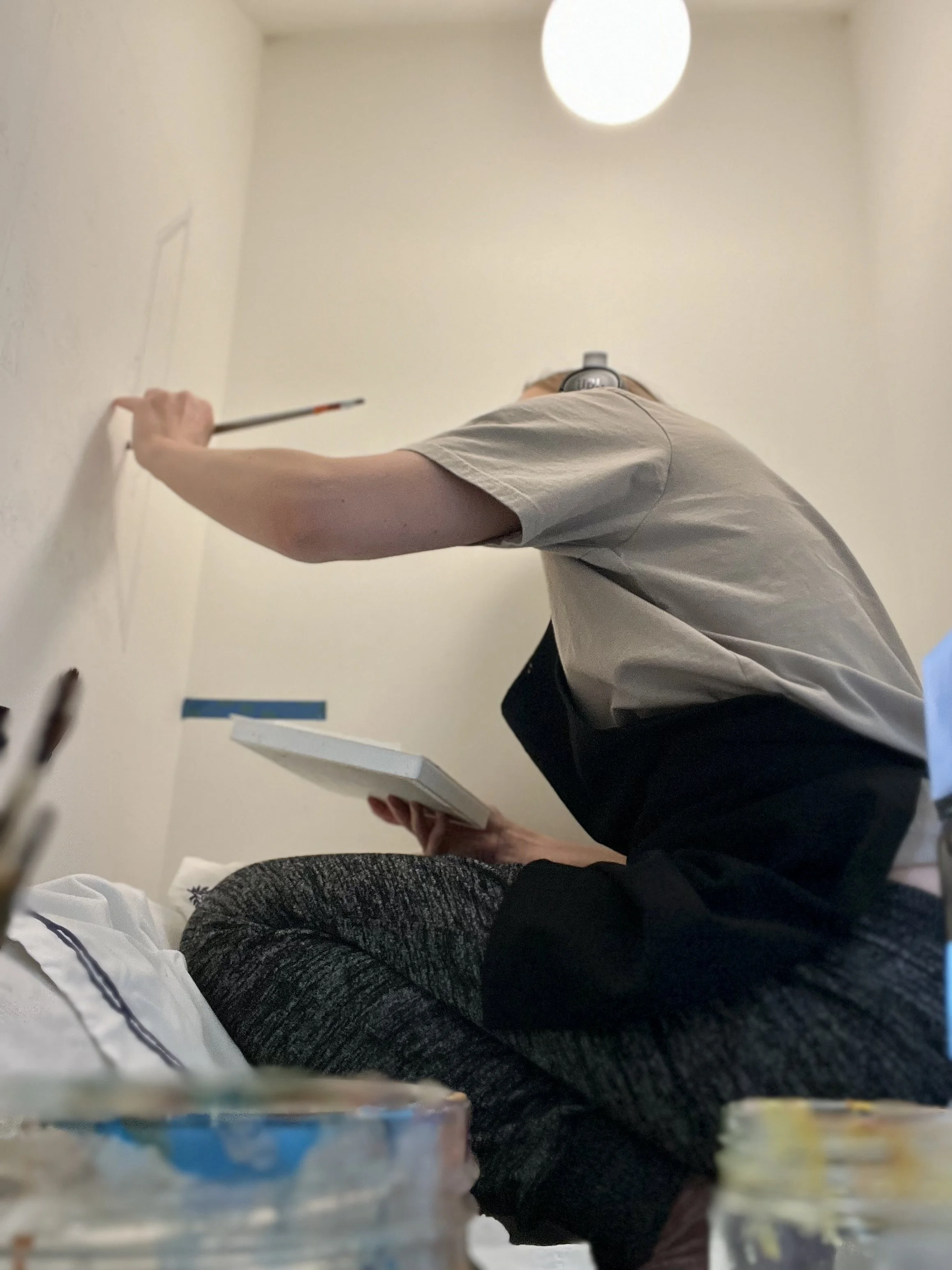 Person sitting cross-legged on a bed, painting or drawing on a wall with a small brush, holding a sketchbook, with art supplies in the foreground and a ceiling light overhead.