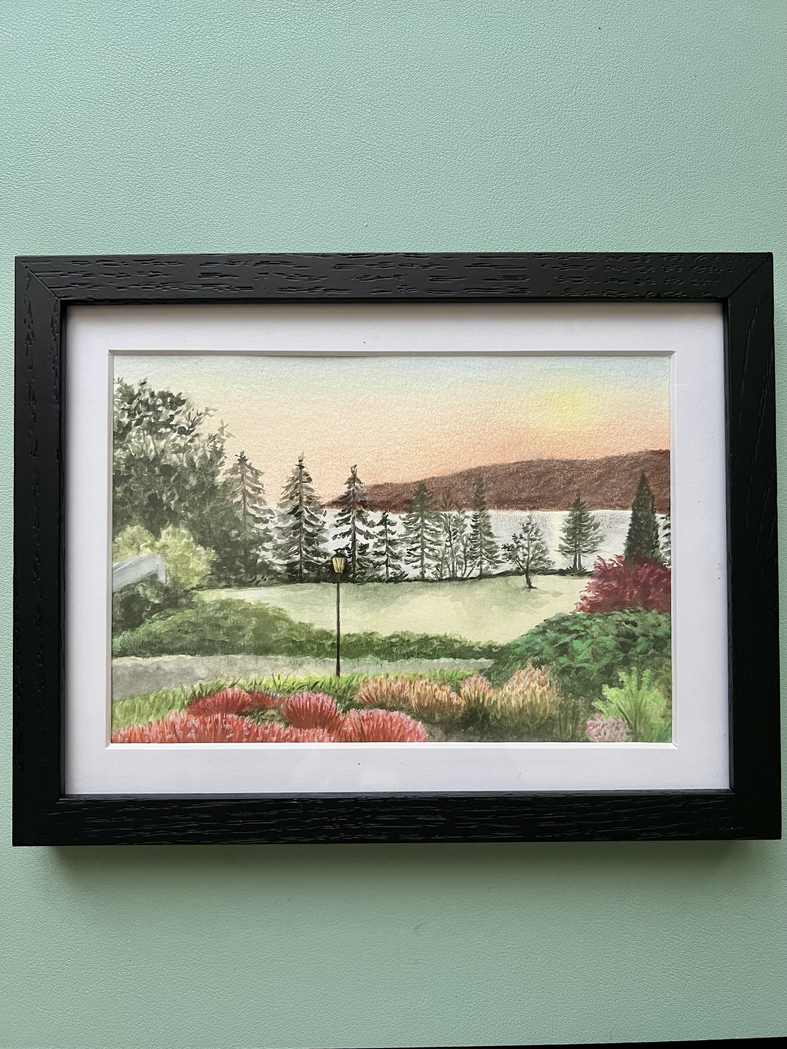 Watercolor painting of a scenic lakeside landscape with trees, a grassy area, colorful bushes, a pathway with a lamppost, and a calm lake with forested hills in the background, during sunset.