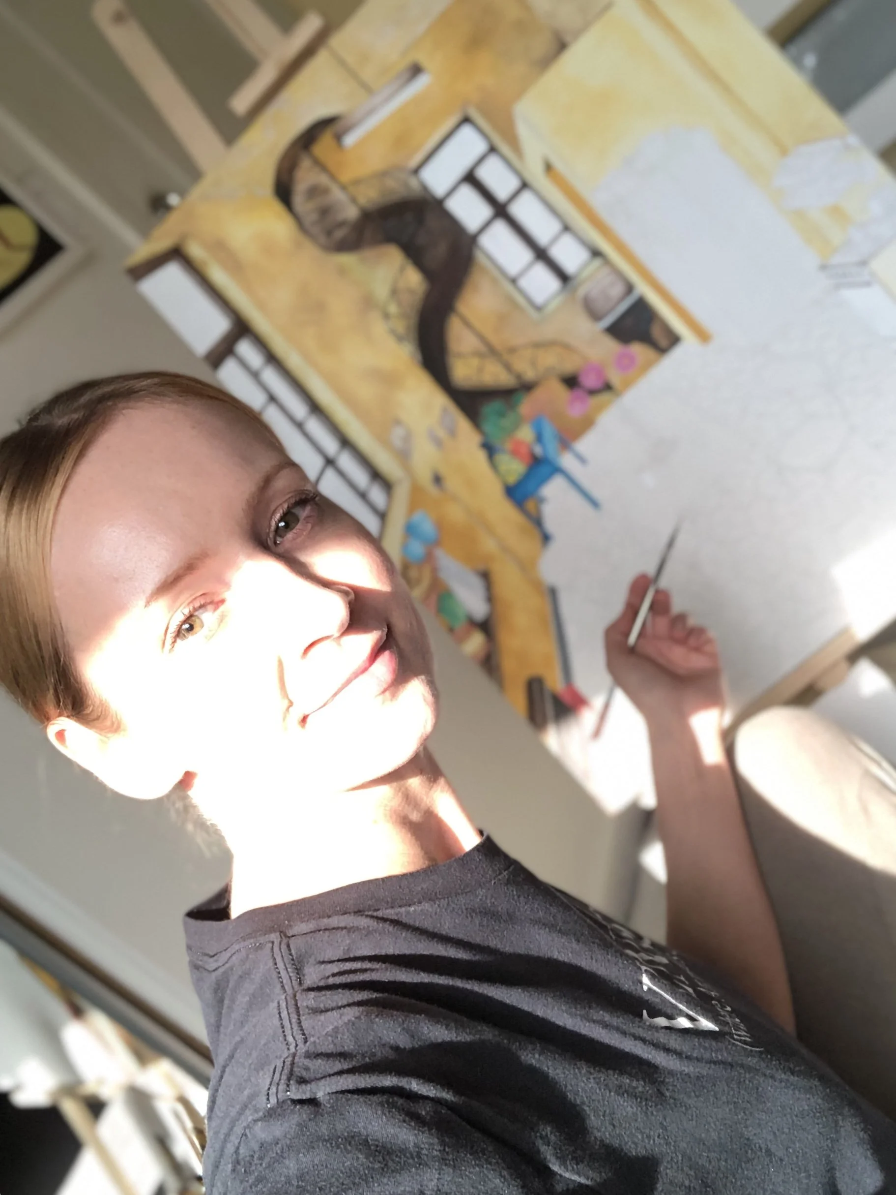 A person with light skin and red hair smiling at the camera, working on a colorful abstract painting in a studio or craft space.