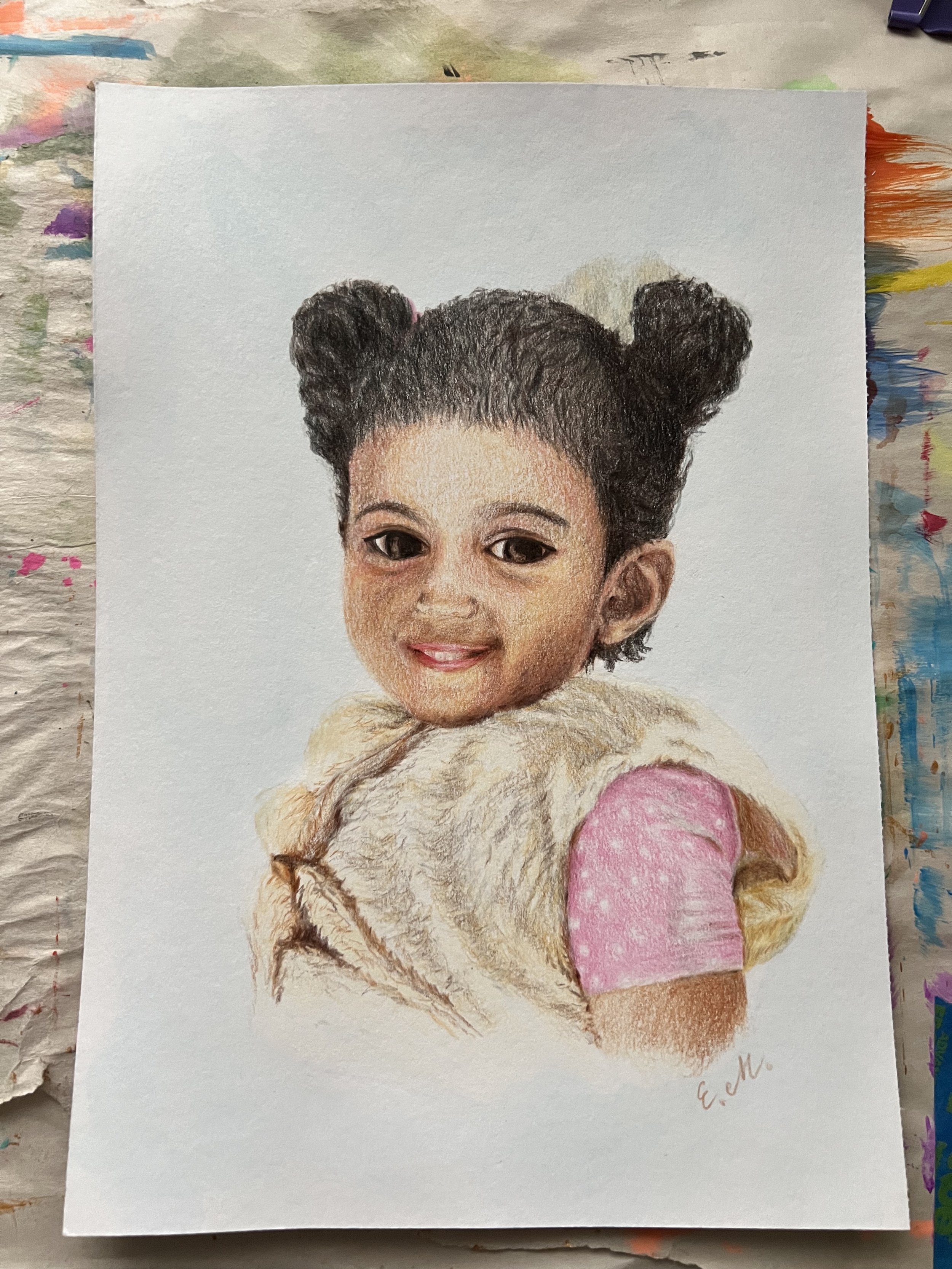 A realistic colored pencil drawing of a young girl with dark hair styled in two buns, smiling, wearing a pink polka dot shirt and a beige furry vest.