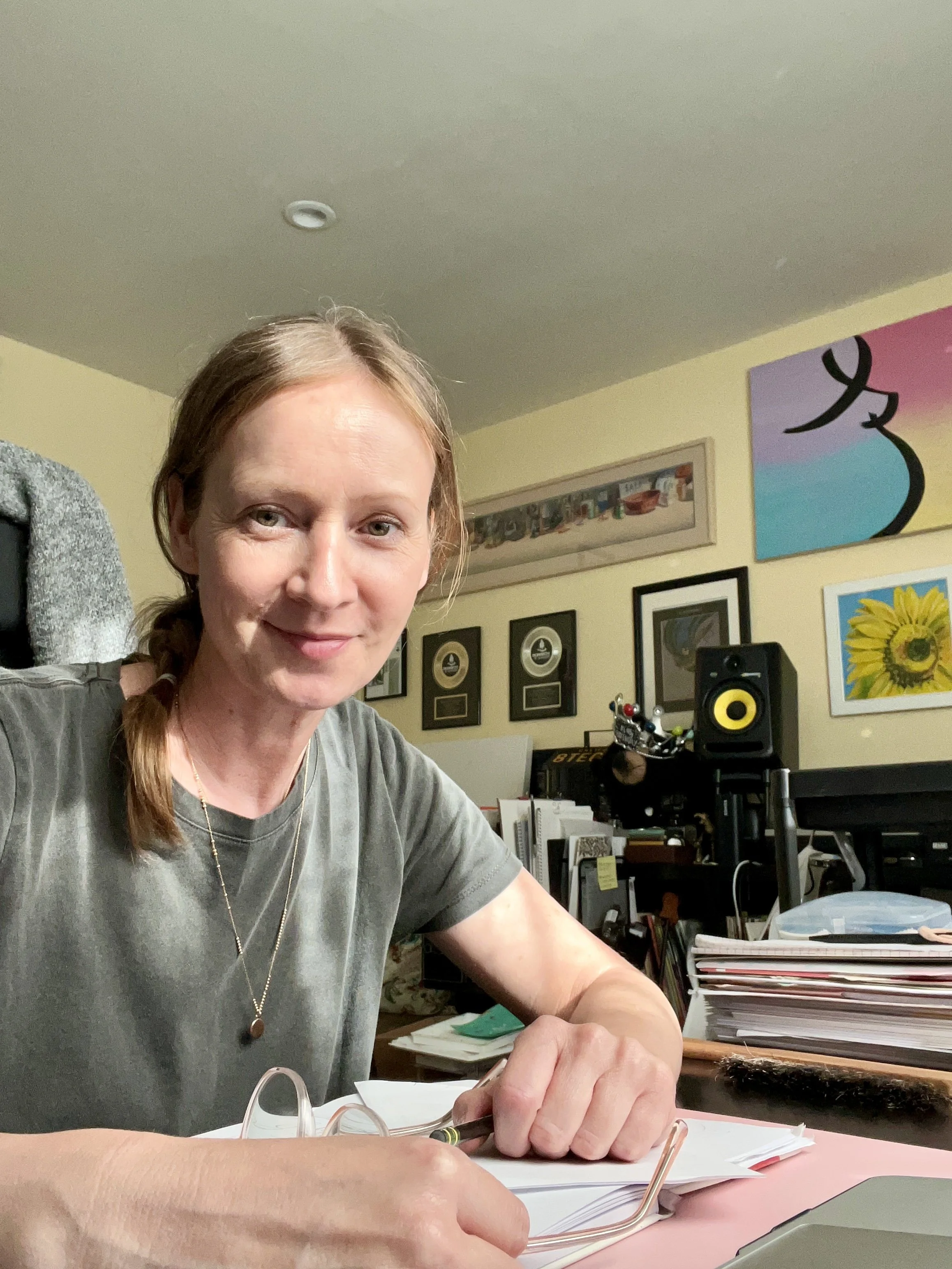 A woman with light brown hair in a braid is sitting at a desk, smiling at the camera. She is holding a pen over a binder or notebook, with stacks of papers and books on her desk. Behind her are framed records, artwork, and audio equipment on the wall.