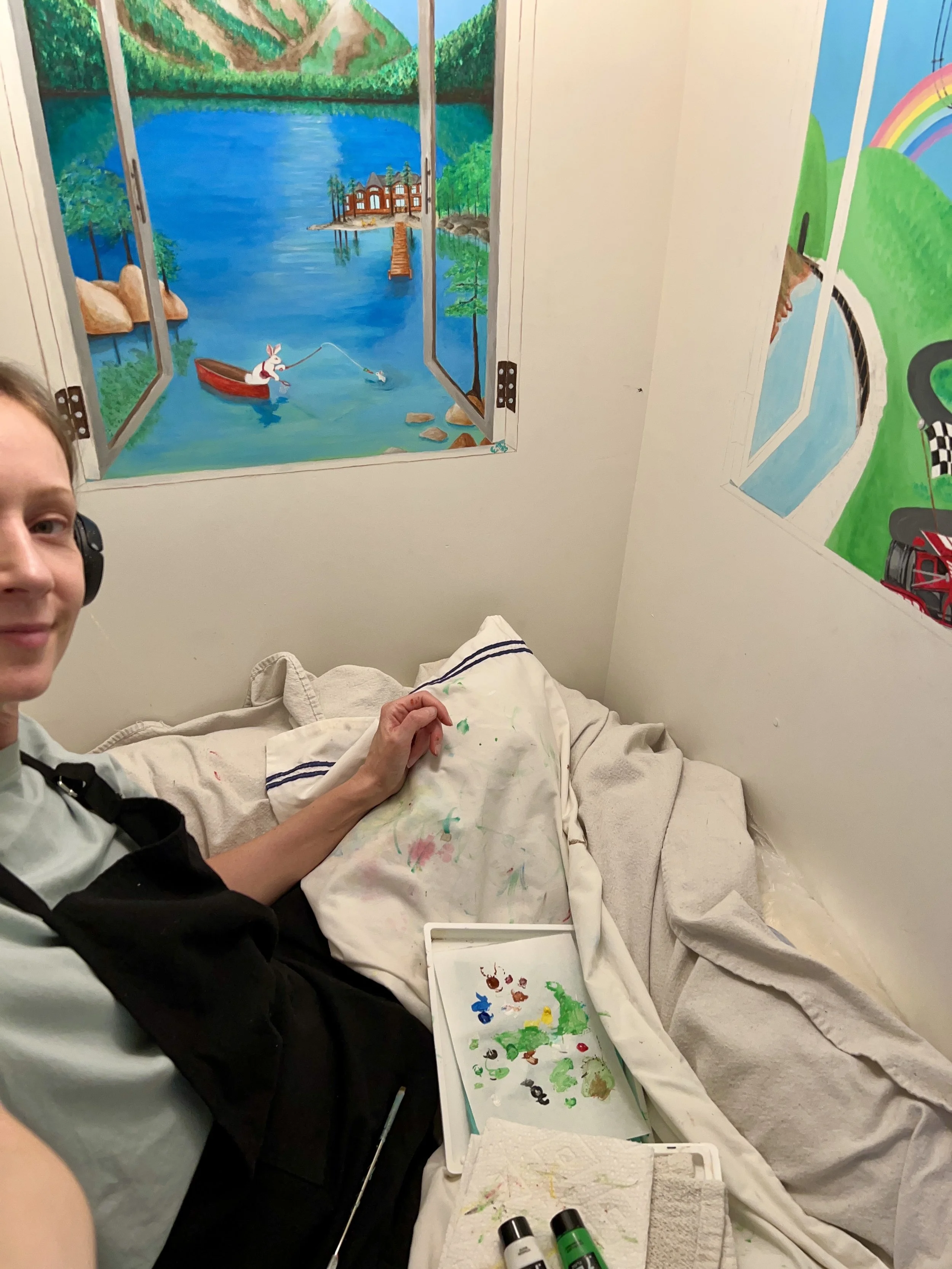 A person painting ocean-themed murals on the walls of a room, with painting supplies and a partially painted scene of a lake, mountains, trees, and a rabbit in a boat.