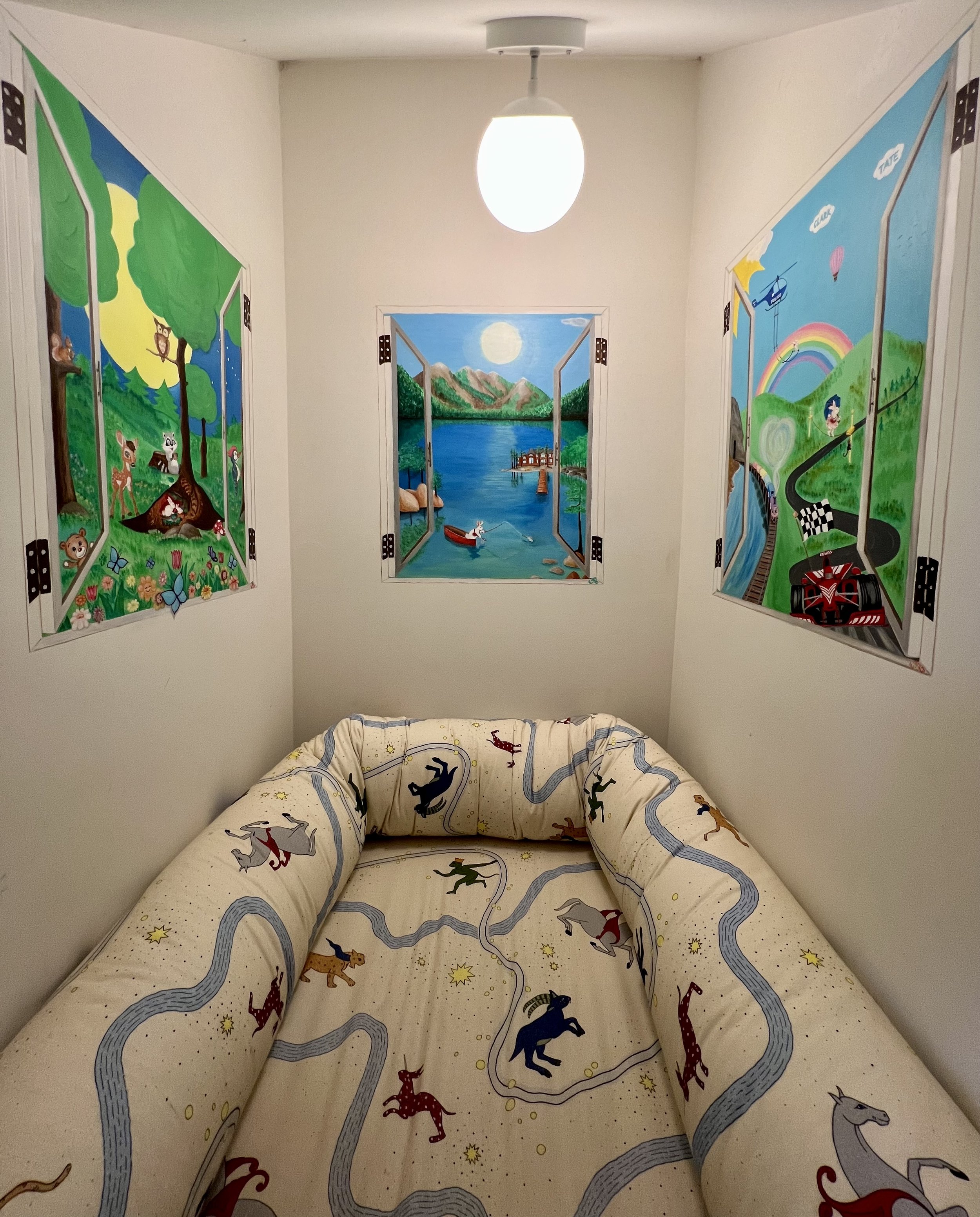 Room with three colorful painted windows depicting outdoor nature scenes and a bed with a kids' bedding featuring animals and winding roads.