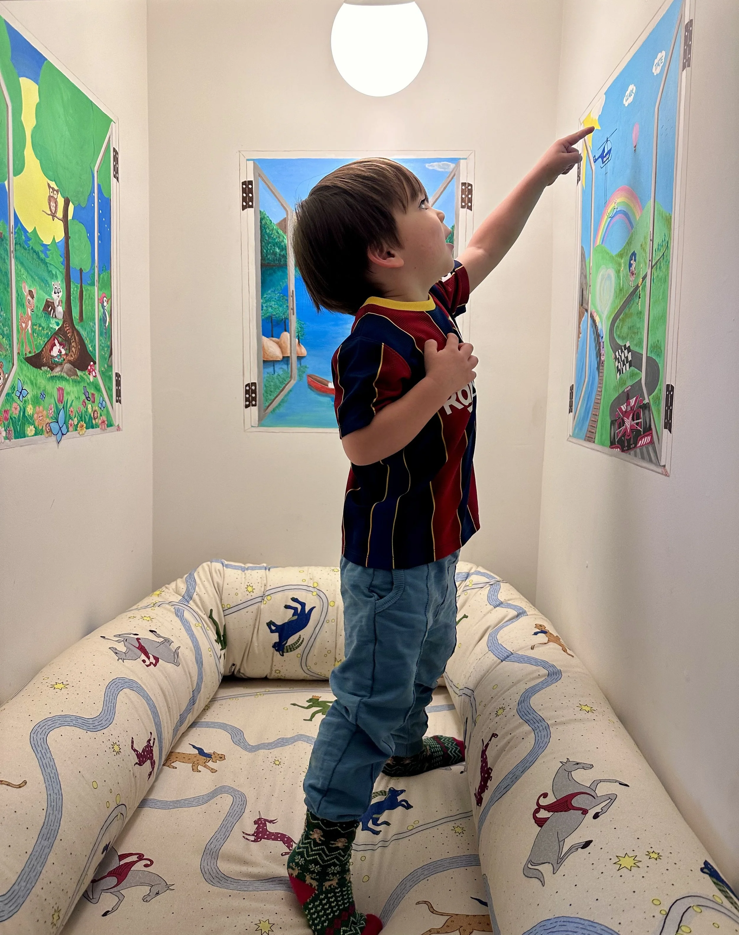 A young boy standing on a bed, reaching up to point at a colorful wall mural depicting a landscape with a rainbow, train, and children playing outdoors.