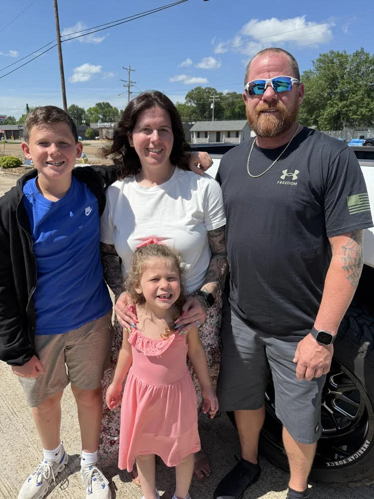 A family of five smiling outdoors on a sunny day, with power lines and a building in the background.