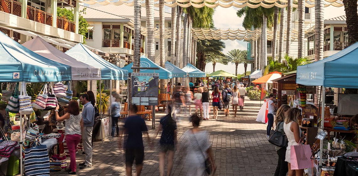 Outdoor marketplace with vendor stalls under tents on a sunny day in a tropical setting, with palm trees and folks browsing and shopping.