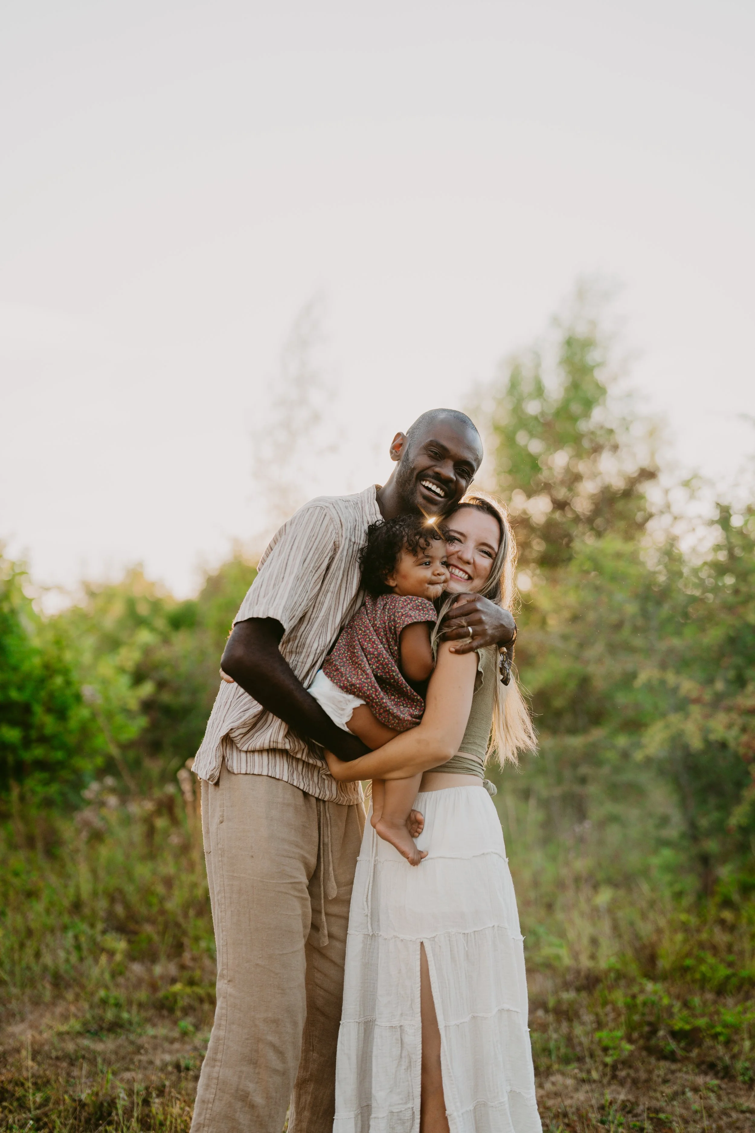 A diverse family group smiling and hugging outdoors during sunset with trees in the background.
