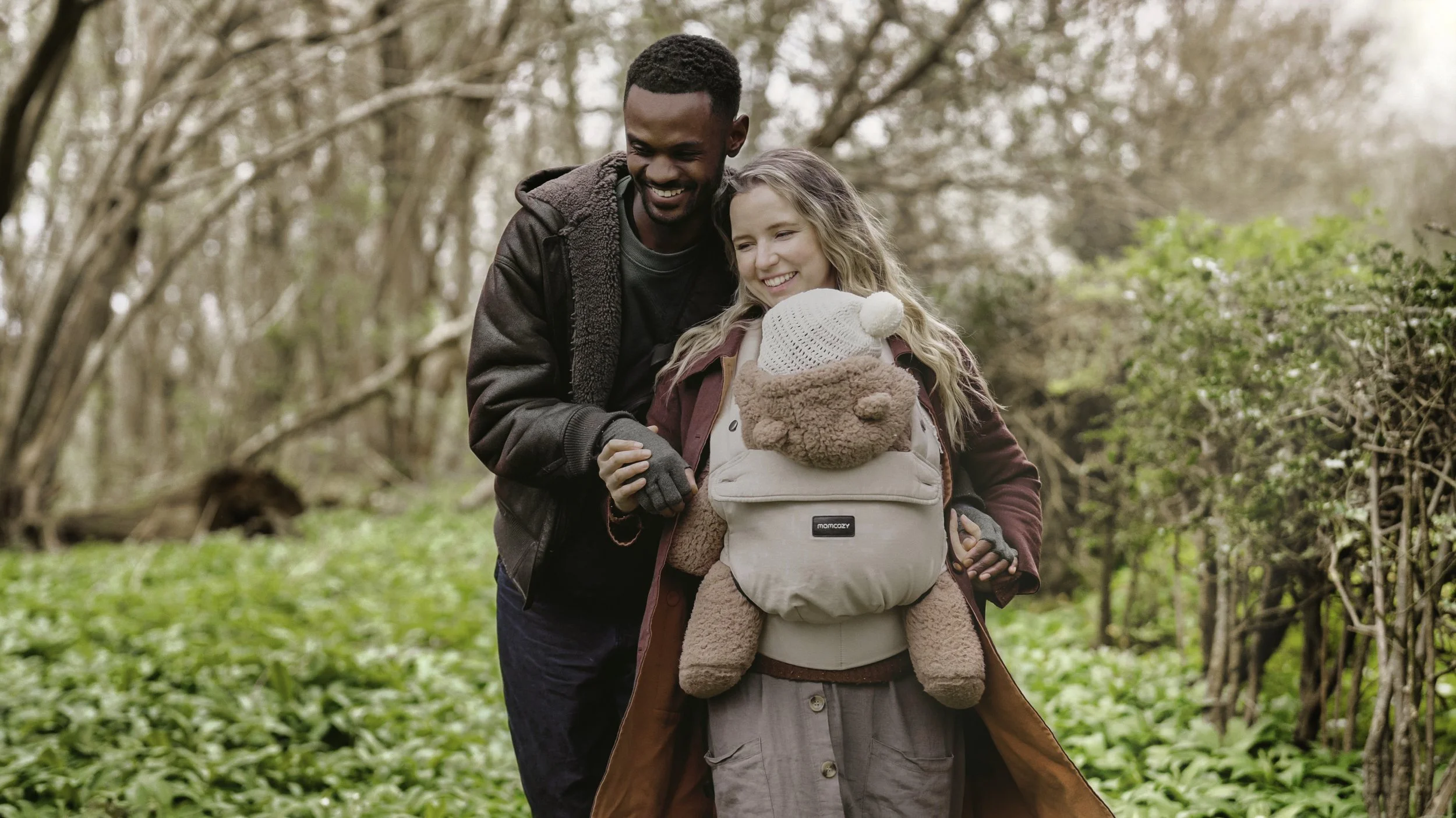 A man and woman walk through a wooded area with a young child in a backpack carrier. The man is smiling and wearing a dark jacket, and the woman is also smiling with a teddy bear hat on her child. The woman is dressed in a coat and beige skirt, and the child’s backpack has a teddy bear face on the front.