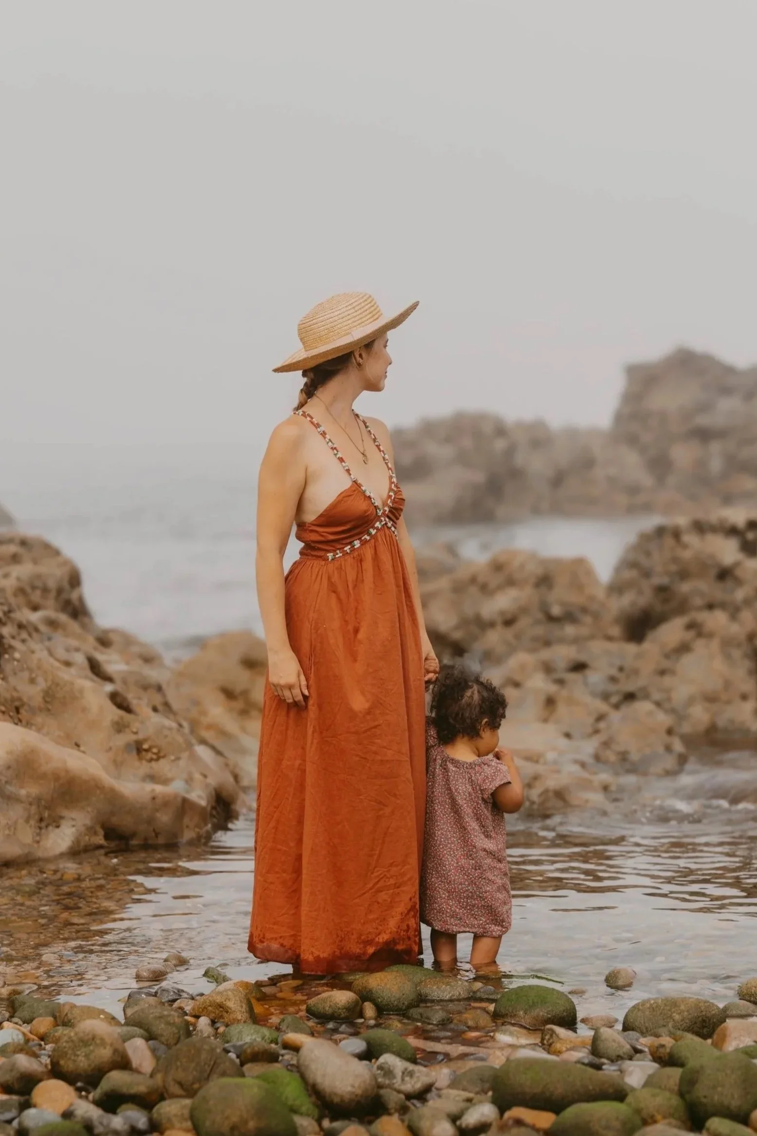 A woman with a hat and a girl in a dress standing in shallow water on a rocky beach, looking out at the ocean with rocks in the background.