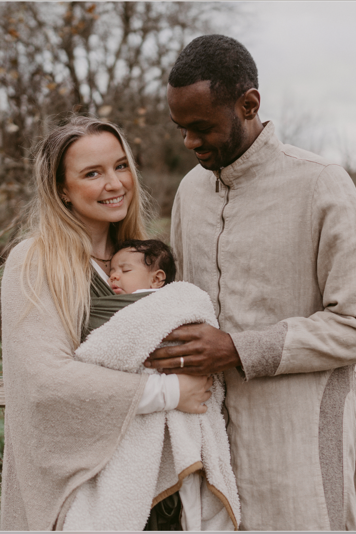A family outdoors, with a woman holding a sleeping baby wrapped in a blanket, a man standing beside them looking at the baby, trees in the background, overcast sky.