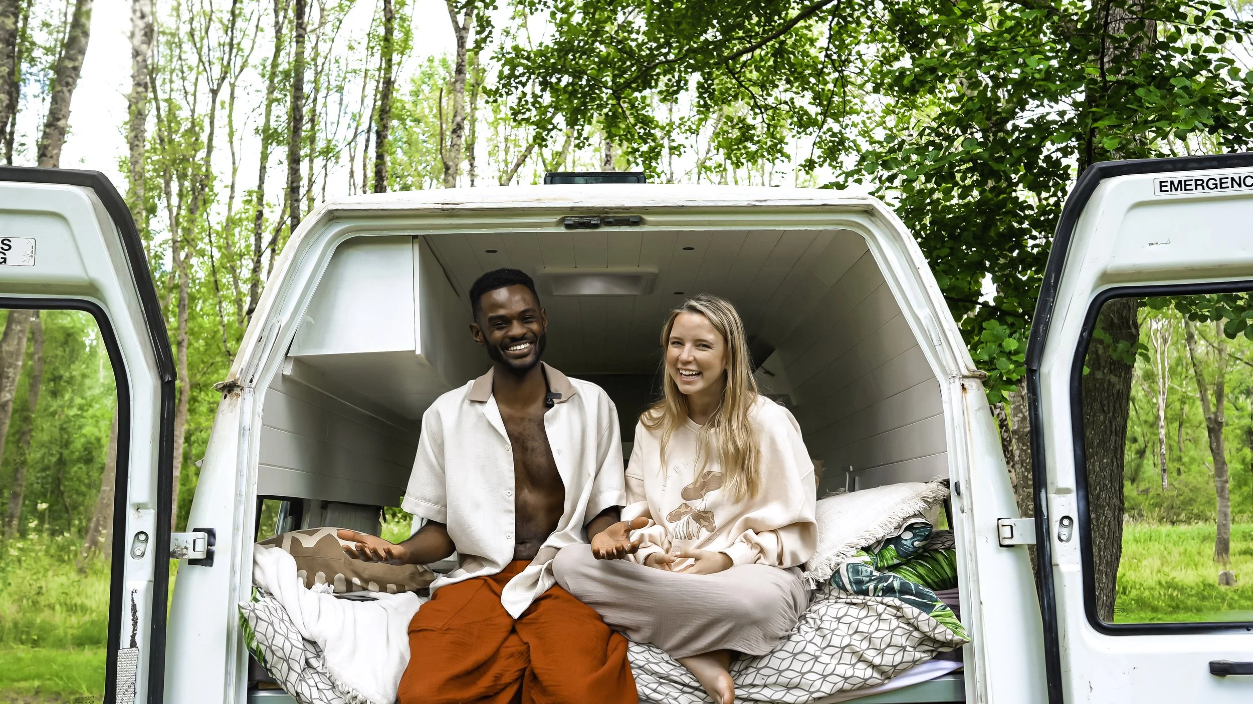 Two smiling young adults, a man and a woman, sitting in the back of a van with the doors open, in a green forest setting.