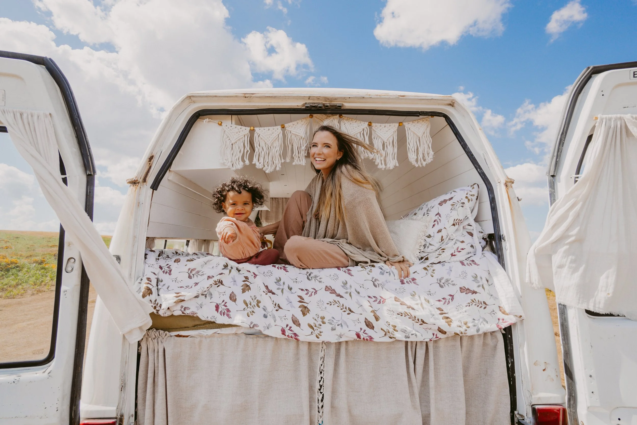 A woman and a young girl sitting inside a decorated vintage camper with open doors, under a blue sky with clouds. The interior has macramé wall hangings and floral bedding.