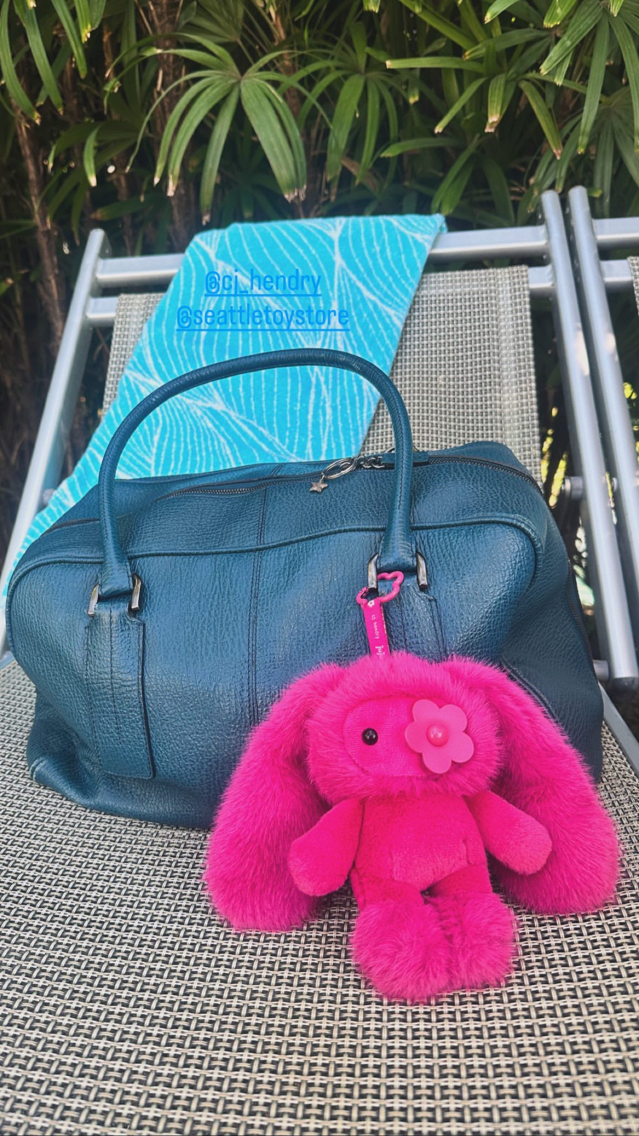 Outdoor photo of a bright pink Juju plush bag charm keychain hanging from a teal leather handbag on a lounge chair, with tropical plants behind
