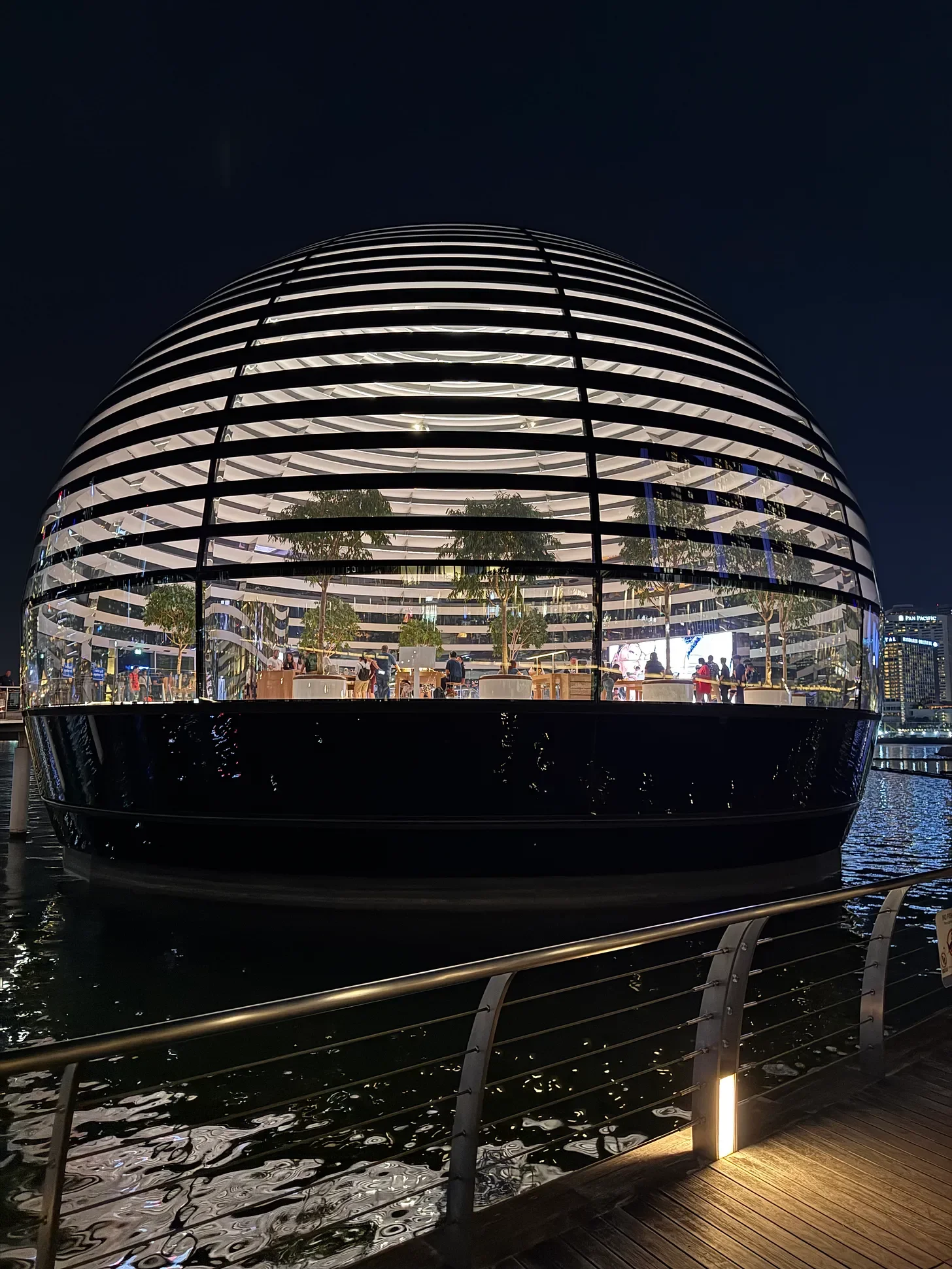 A brightly lit, spherical Apple Store sits on the water outside Marina Bay Sands in Singapore, its glass-and-slat facade revealing indoor trees and shoppers; a waterfront railing runs across the foreground.