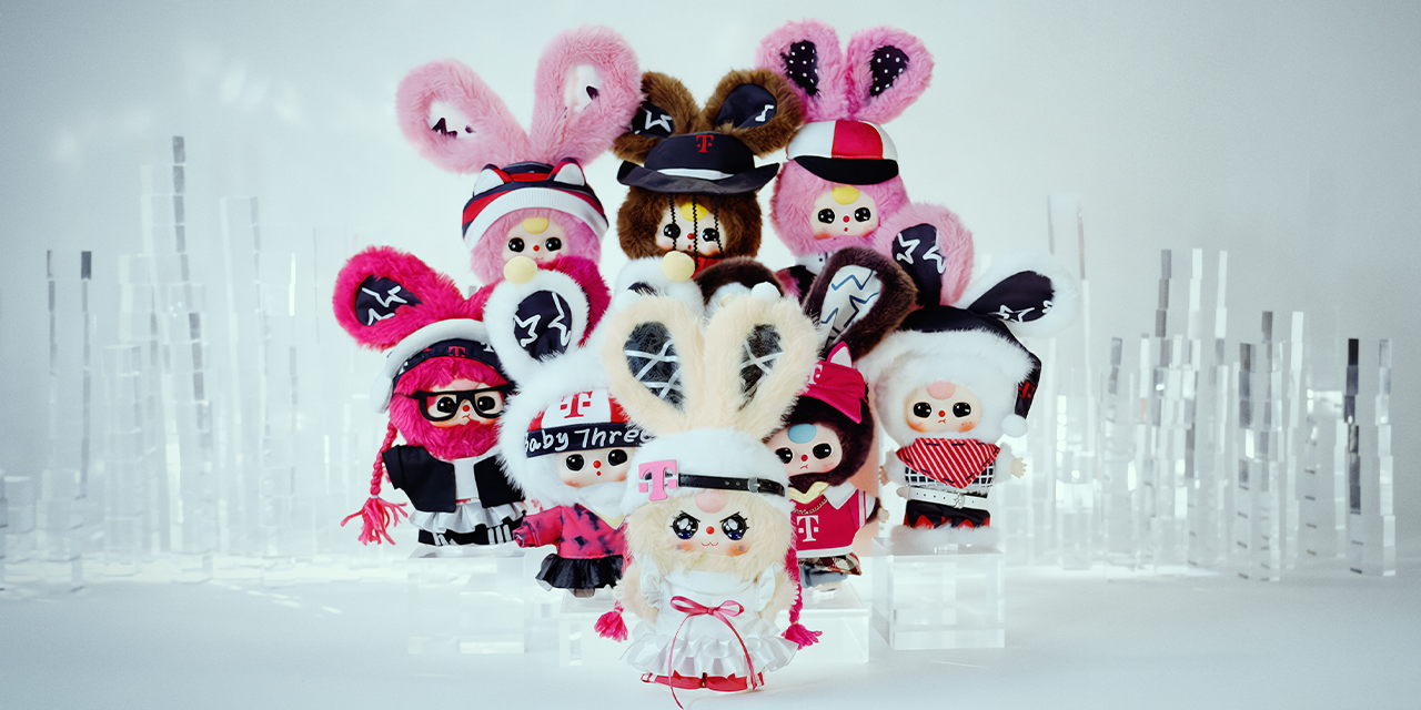 Lineup of nine Baby Three plush dolls arranged in rows against a white backdrop. Each bunny-eared character wears T-Mobile–inspired outfits in pink, white, black, and red: hoodies, skirts, caps, and headbands with the T logo and star graphics.