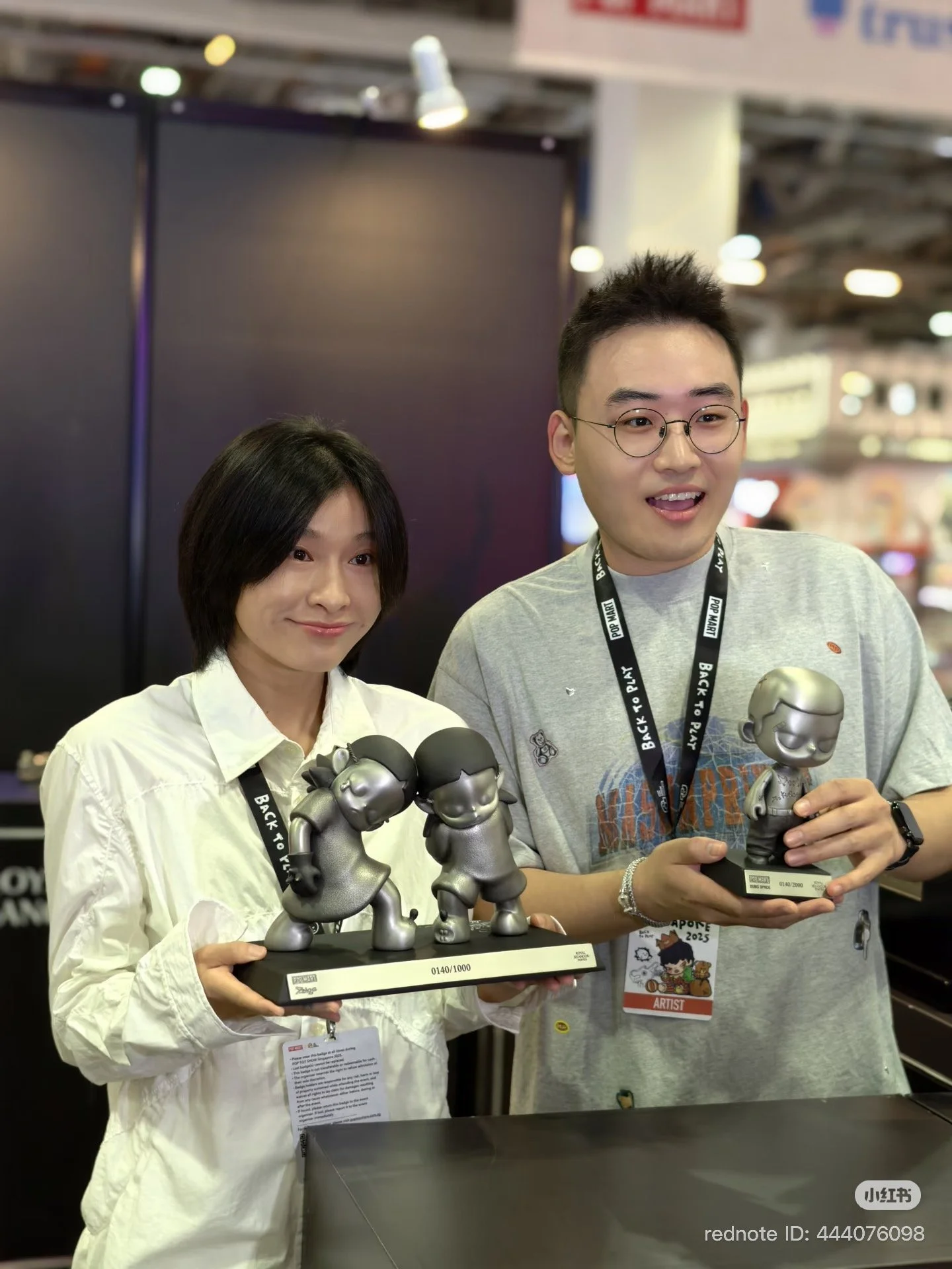Screenshot of two people at an expo booth holding limited-edition pewter character sculptures. Both wear event lanyards, and a caption below references a Royal Selangor × POP MART pewter showcase for “KUBO: TIN ME 200%” and “Zsiga.”