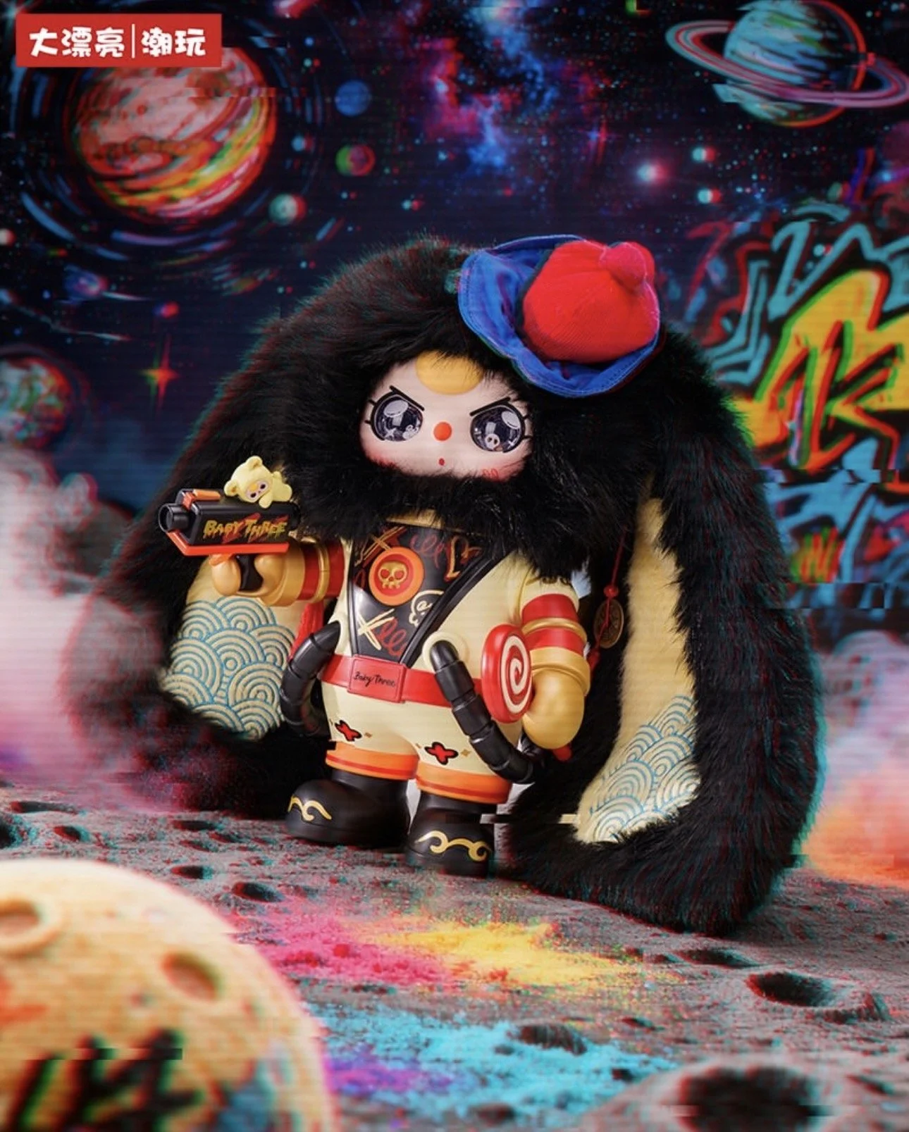 Promotional image of a Baby Three Dreamer character wearing a black furry bunny-hood cape over a beige-and-gold space suit, holding a small toy blaster and standing on a cratered, rainbow-splashed moon surface