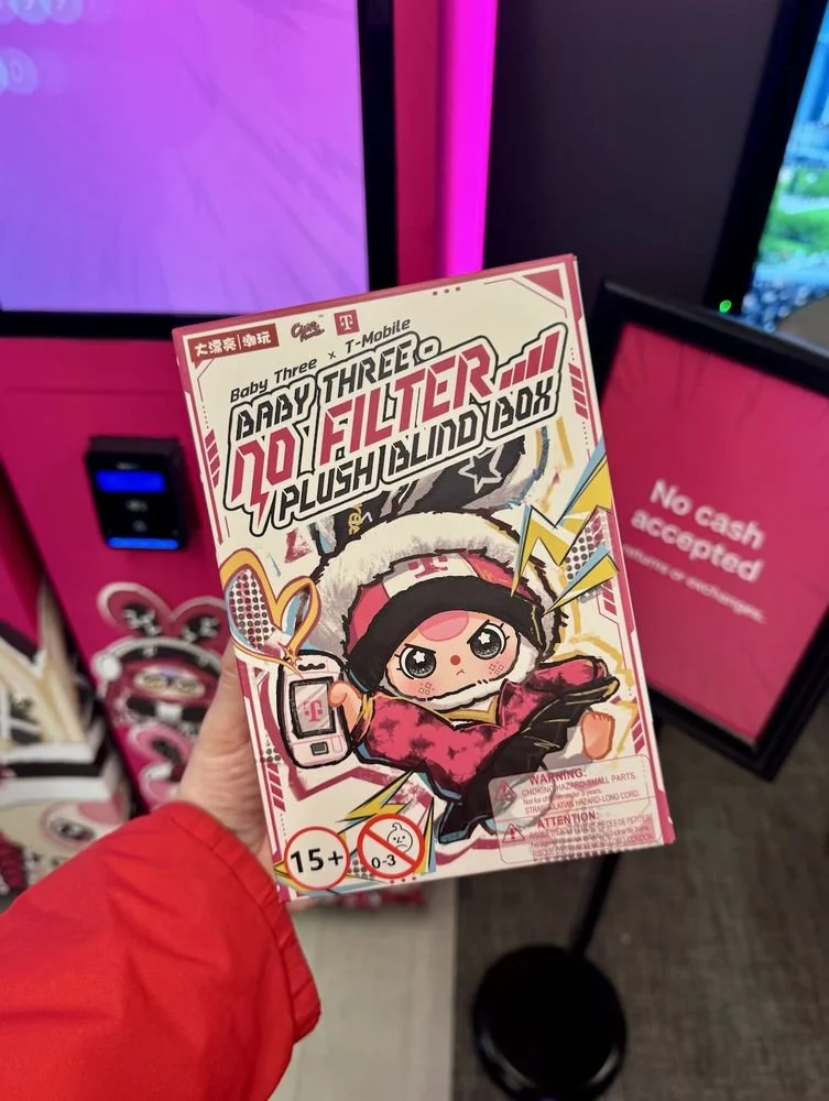 A hand holding a Baby Three x T-Mobile “No Filter” plush blind box in front of a bright pink display and a “No cash accepted” sign.