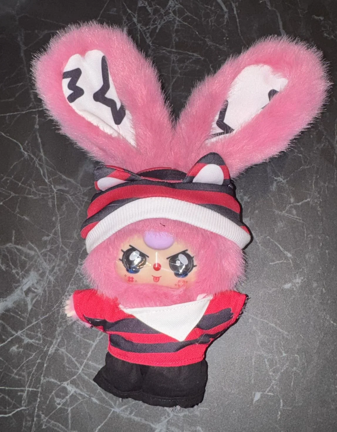 Baby Three Zing plush with pink fur and tall ears wearing a red-and-black striped outfit and a headwrap-style hat on a dark surface.