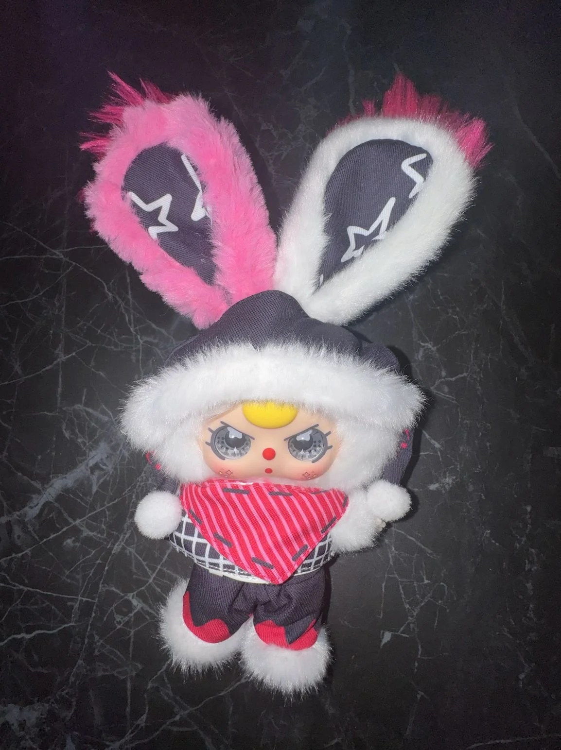 Baby Three Luxe plush on a dark surface wearing a hood with white faux fur trim and a red patterned bandana, with one pink ear and one white ear.