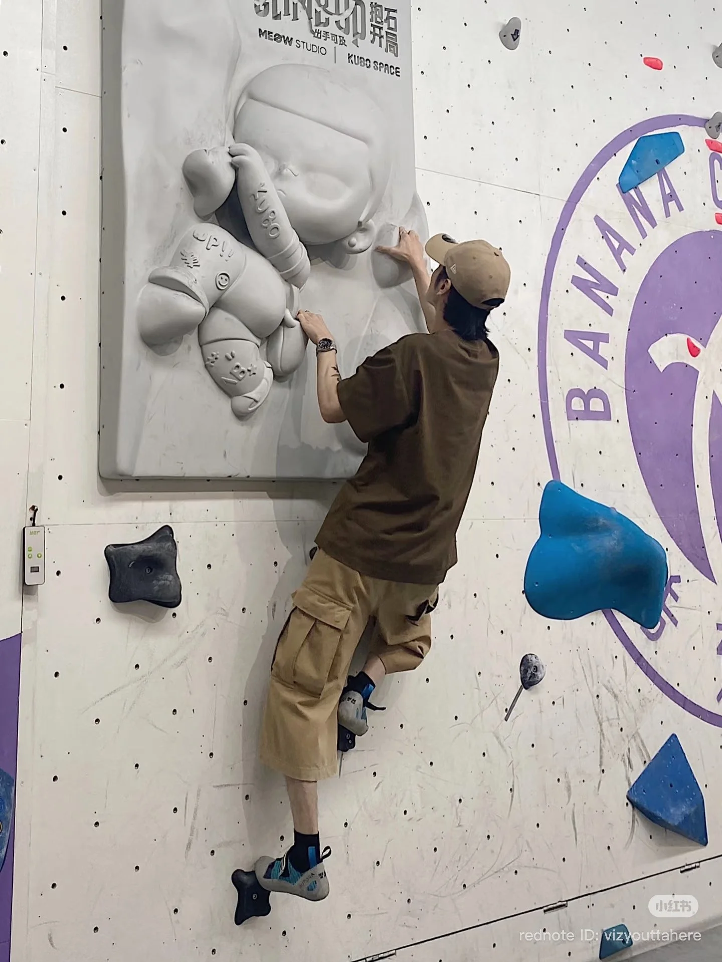 A climber tests a bouldering wall featuring an oversized KUBO “CLIMB UP” relief panel, using the sculpted “rock” forms as part of the route—an on-the-wall crossover of character IP and climbing.