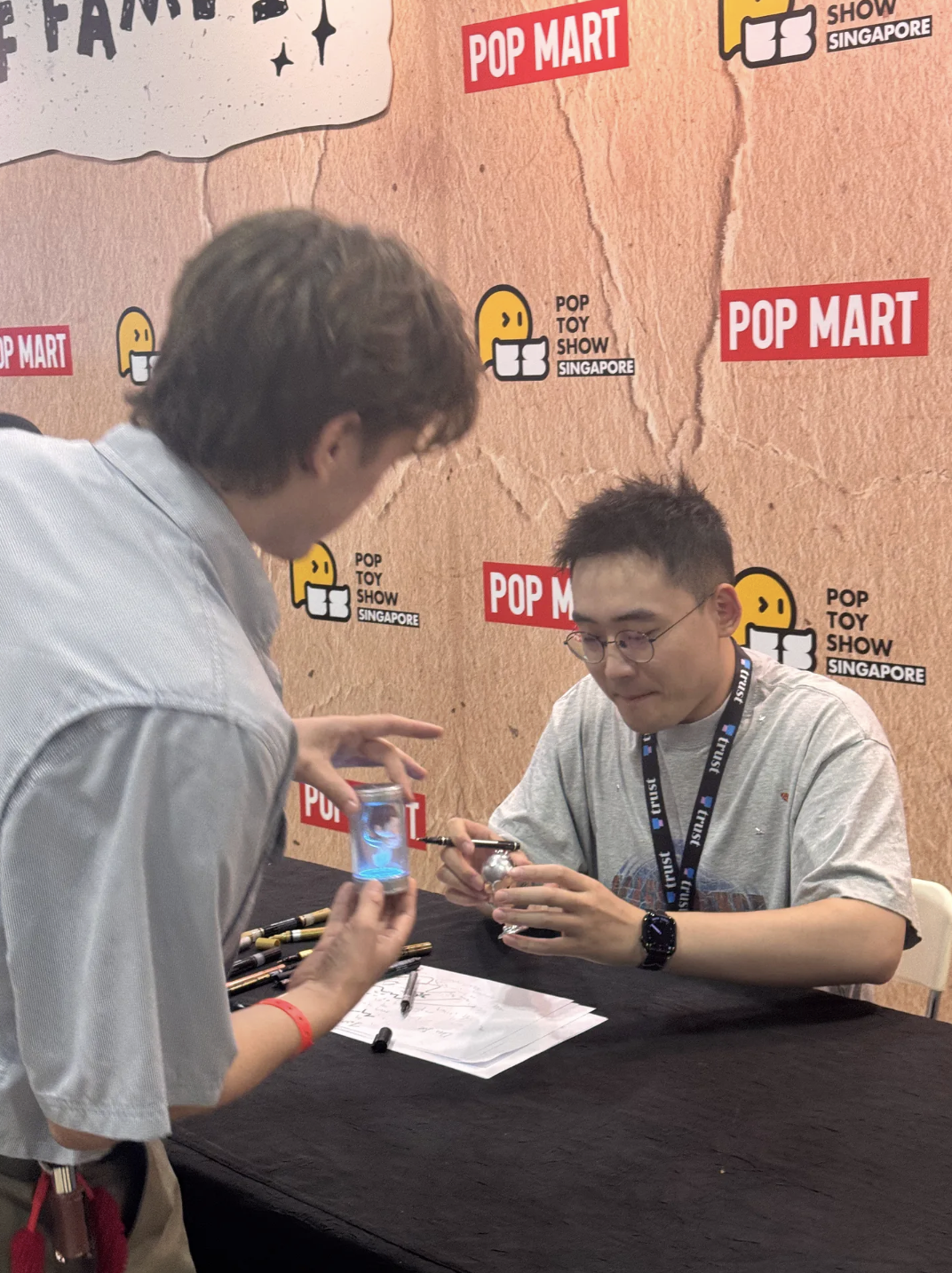In front of a POP MART / “Pop Toy Show Singapore” photo wall, Bao sits at a signing table holding a small figure while I lean in holding a clear capsule with a blue-lit figure inside.