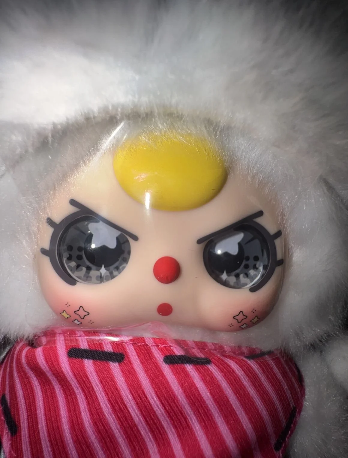 Close-up of Baby Three Luxe face with marbled gray eyes, a yellow forehead detail, cheek sparkles, and clear protective plastic film covering the faceplate.