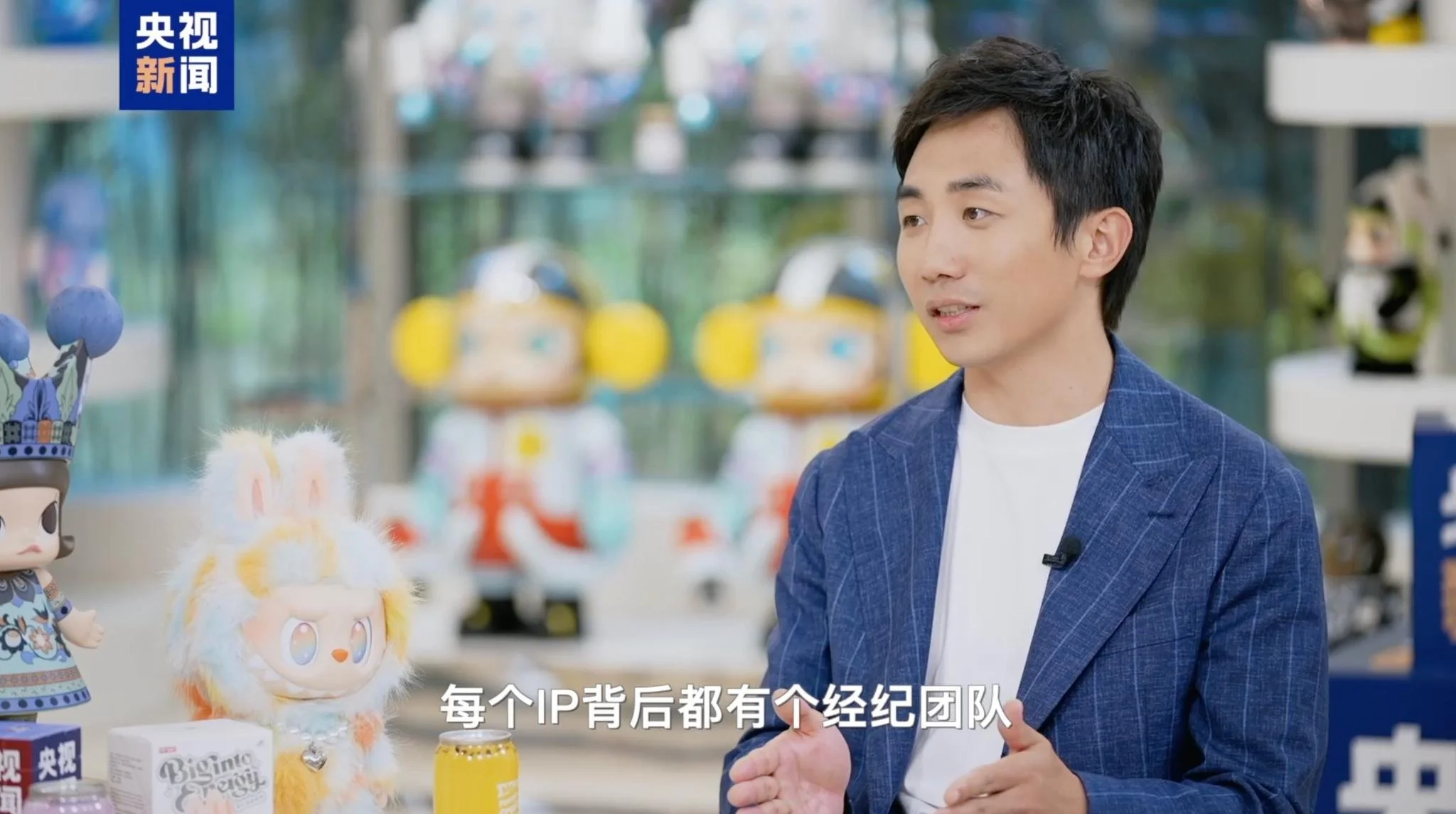 A young man in a blue blazer speaks during a CCTV News interview; designer toy figures and Chinese subtitles (“每个 IP 背后都有个经纪团队” — “Every IP has its own management team”) sit in the foreground. The young man is Pop Mart CEO Wang Ning.