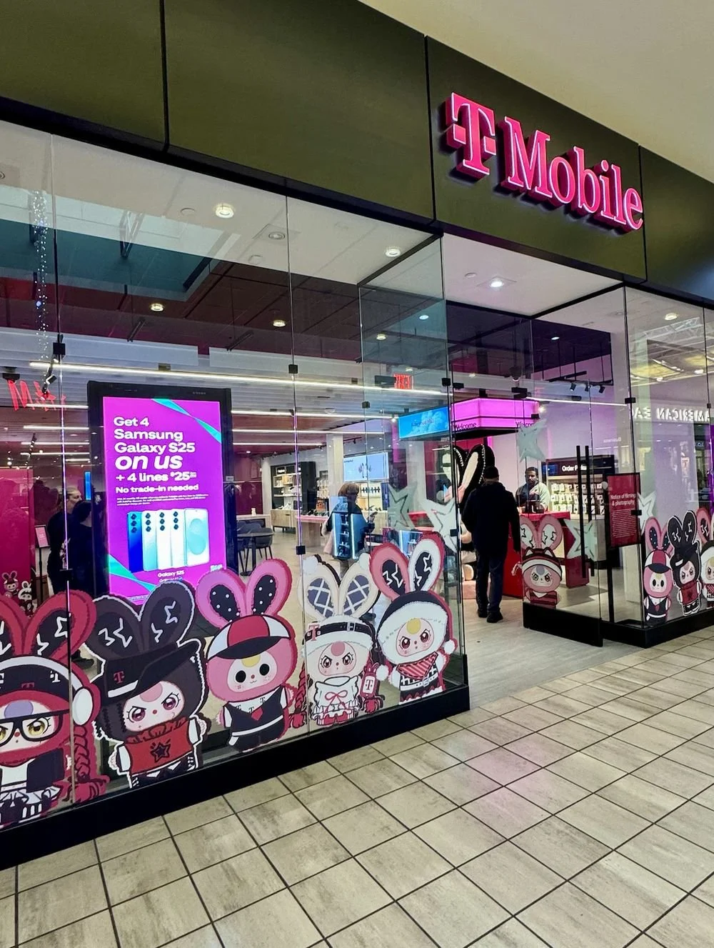 Glass-fronted T-Mobile store in a mall with large Baby Three character decals across the windows and a bright magenta digital display advertising a Samsung Galaxy S25 deal inside.