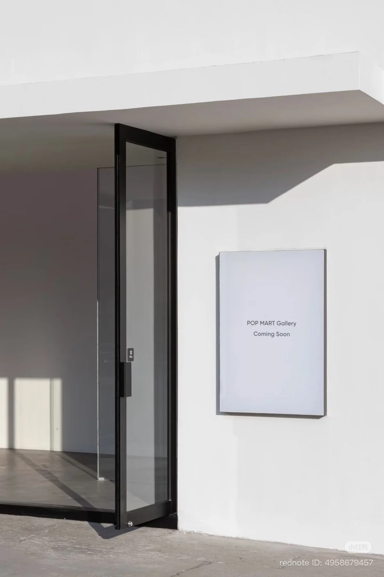 Exterior doorway with a propped-open glass door and a wall-mounted sign reading “POP MART Gallery Coming Soon,” lit by sharp afternoon shadows.