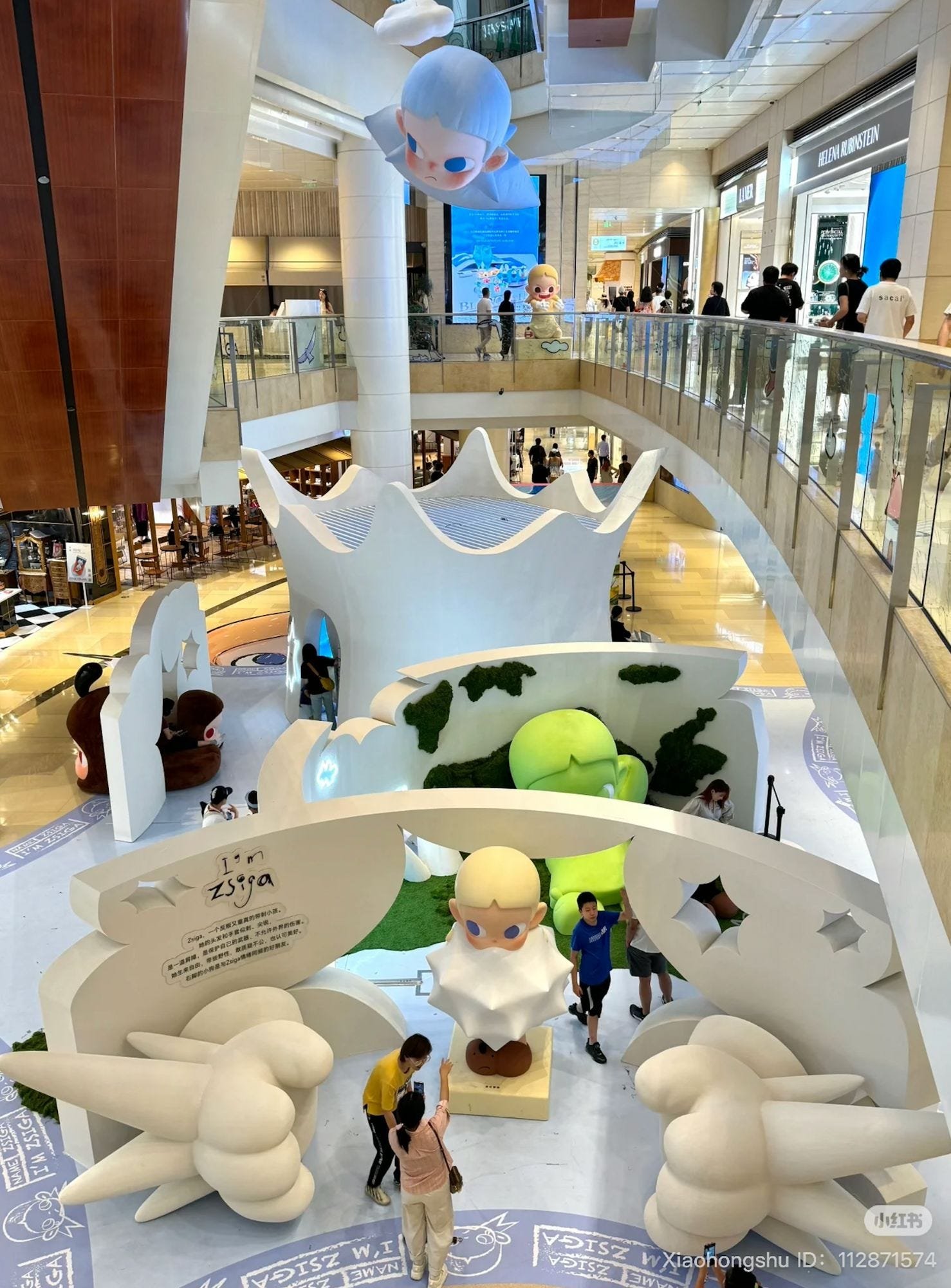Overhead view of a multi-level shopping mall atrium featuring the “I’m Zsiga” installation with multiple giant character figures and sculptural set pieces, as shoppers watch from upper-floor railings.