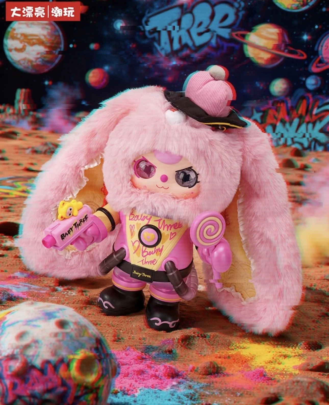Promotional image of a Baby Three Dreamer character wearing a black furry bunny-hood cape over a beige-and-gold space suit, holding a small toy blaster and standing on a cratered, rainbow-splashed moon surface