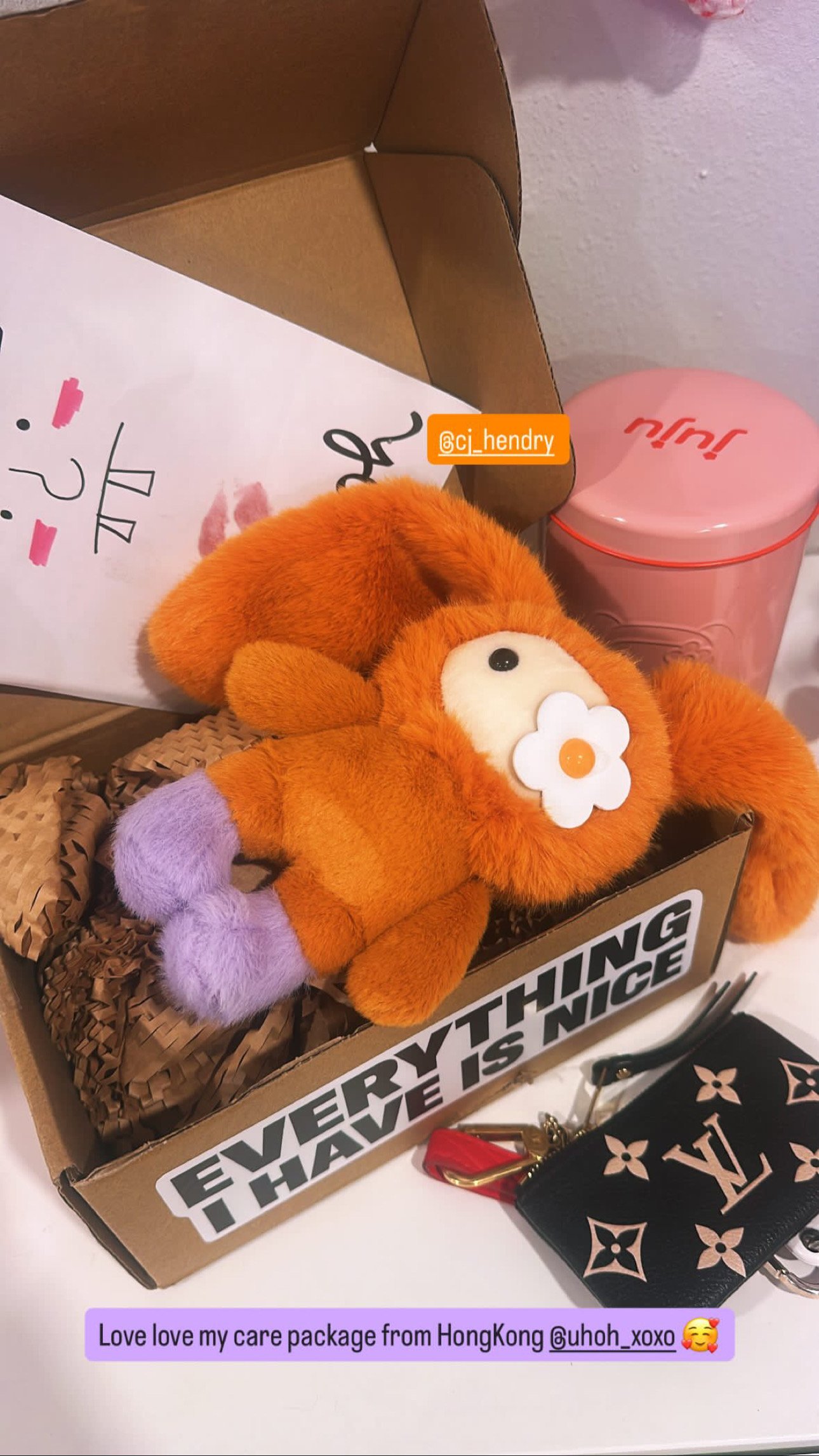 Close-up of an orange Juju plush bag charm keychain with long ears and a white flower detail, lying in an open gift box with crinkle paper.