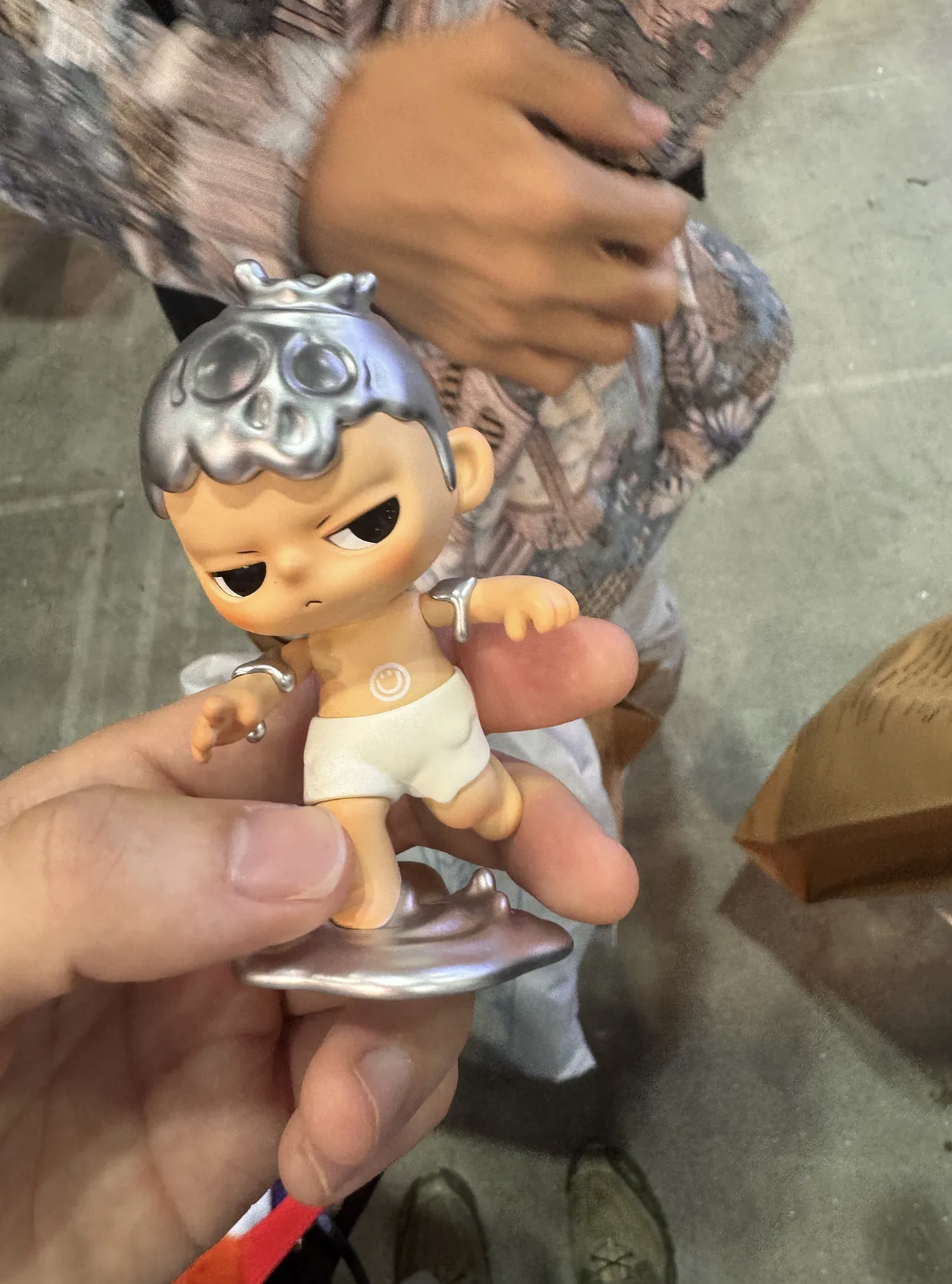 A hand holds a KUBO figure wearing white shorts, posed mid-step on a metallic silver puddle base; the figure has a shiny silver “melting” headpiece and silver drips on the shoulders and arms.