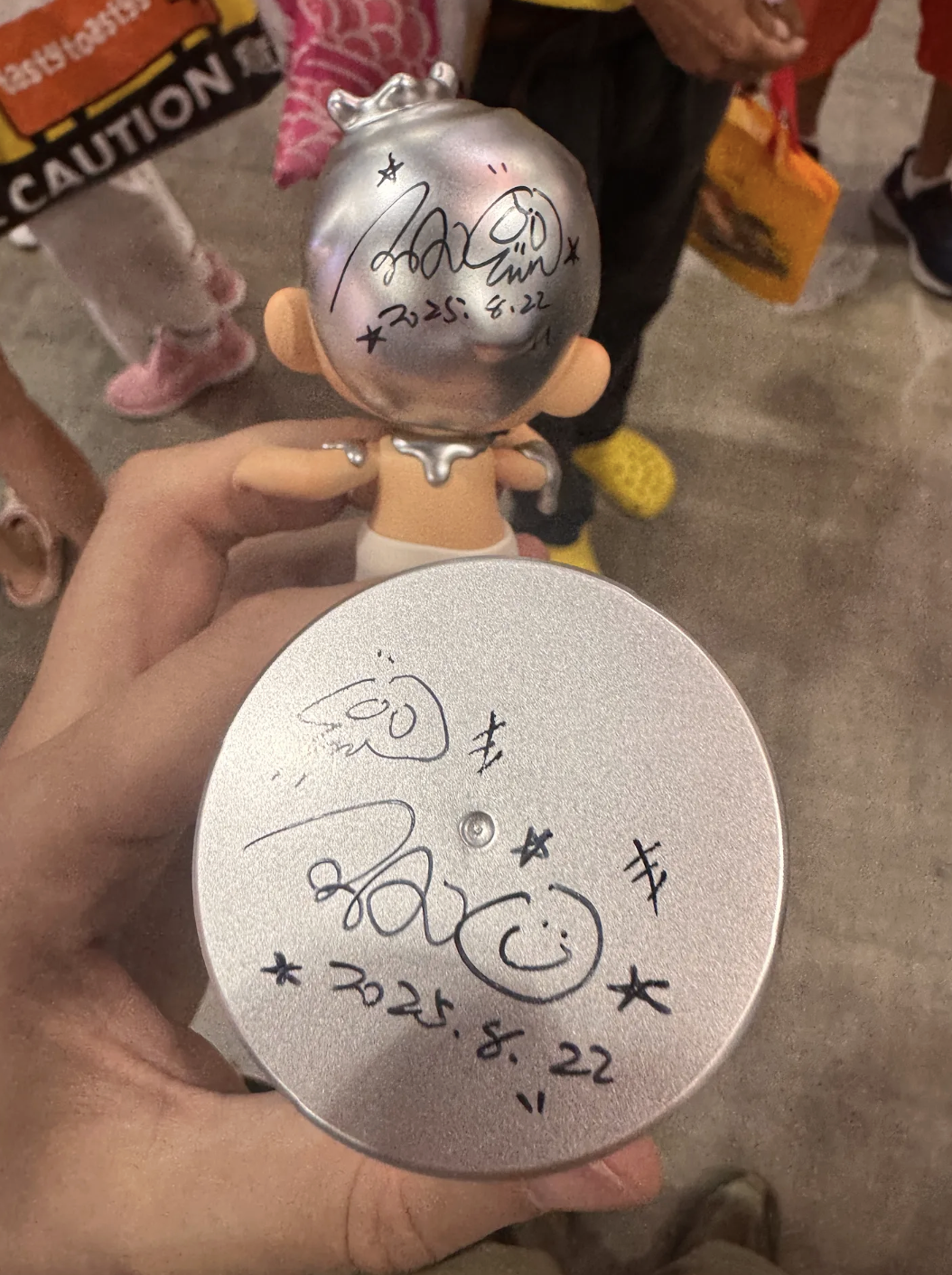 A hand holds the signed KUBO figure (signature on the back of the silver head) while a round metal lid in the foreground shows a black-marker signature, smiley doodle, and the date “2025.8.22.”