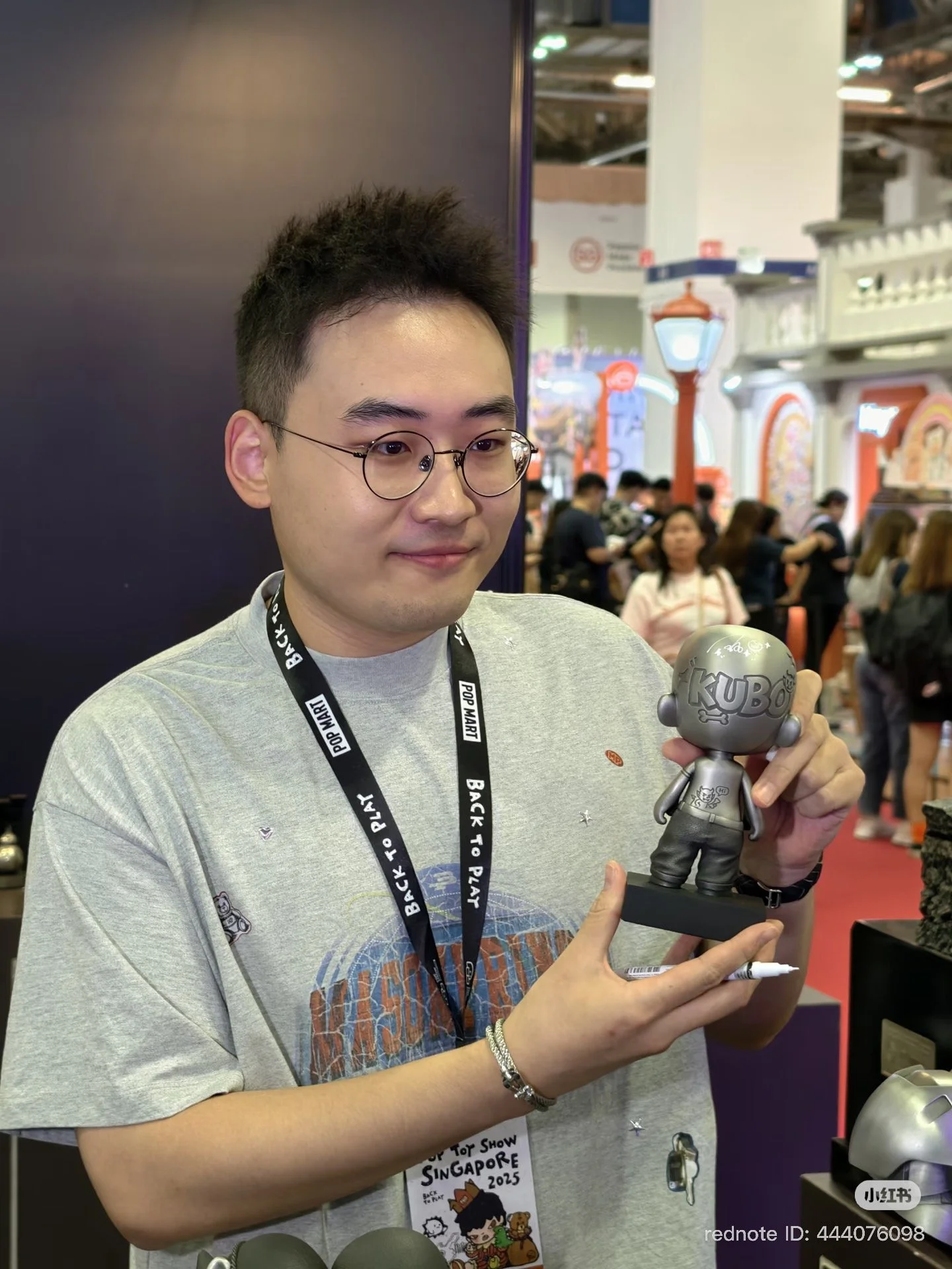 A person wearing glasses and a POP MART “BACK TO PLAY” lanyard holds the pewter KUBO figure on its base at a busy convention booth.