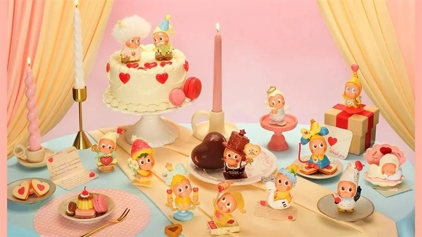 A pink-and-cream Valentine dessert table set: a frosted cake with hearts, pastel candles, cookies, macarons, and chocolate. Small Twinkle Twinkle figures in party hats and cute costumes are arranged across the table, including one lounging on a heart