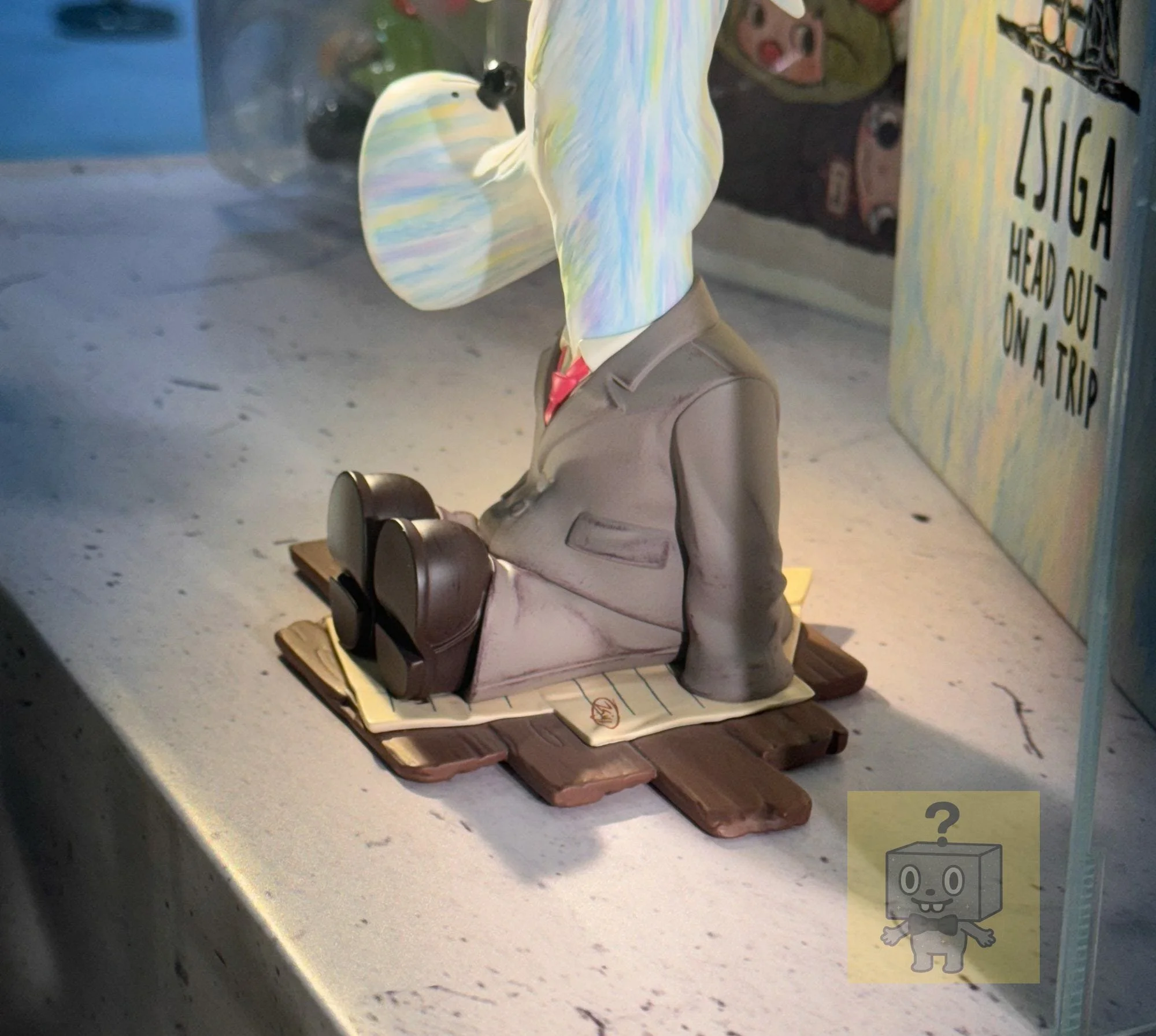 Detail shot of the seated Zsiga figure in a gray suit with black shoes on a layered base resembling wooden boards and paper sheets; the words “ZSIGA HEAD OUT ON A TRIP” are partially visible on packaging in the background.