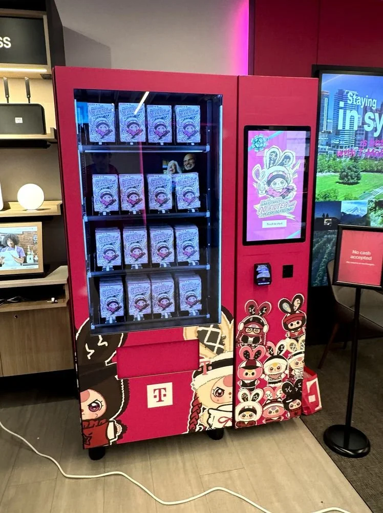 A pink T-Mobile vending machine decorated with Baby Three characters, filled with “No Filter” plush blind boxes and a touchscreen that says “Touch to start.”
