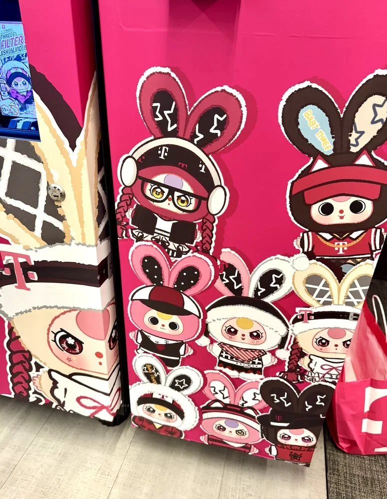 Close-up of Baby Three character graphics on a bright magenta panel inside a T-Mobile store, showing multiple bunny-eared figures in branded outfits.