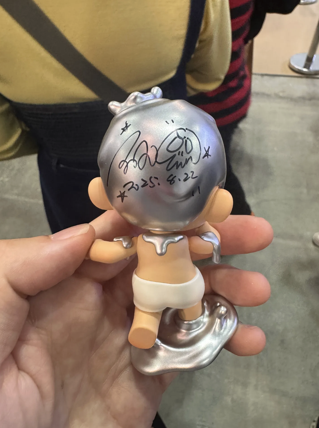 Back view of the same KUBO figure in a hand; the metallic silver head is signed in black marker with small star doodles and the date “2025.8.22.”